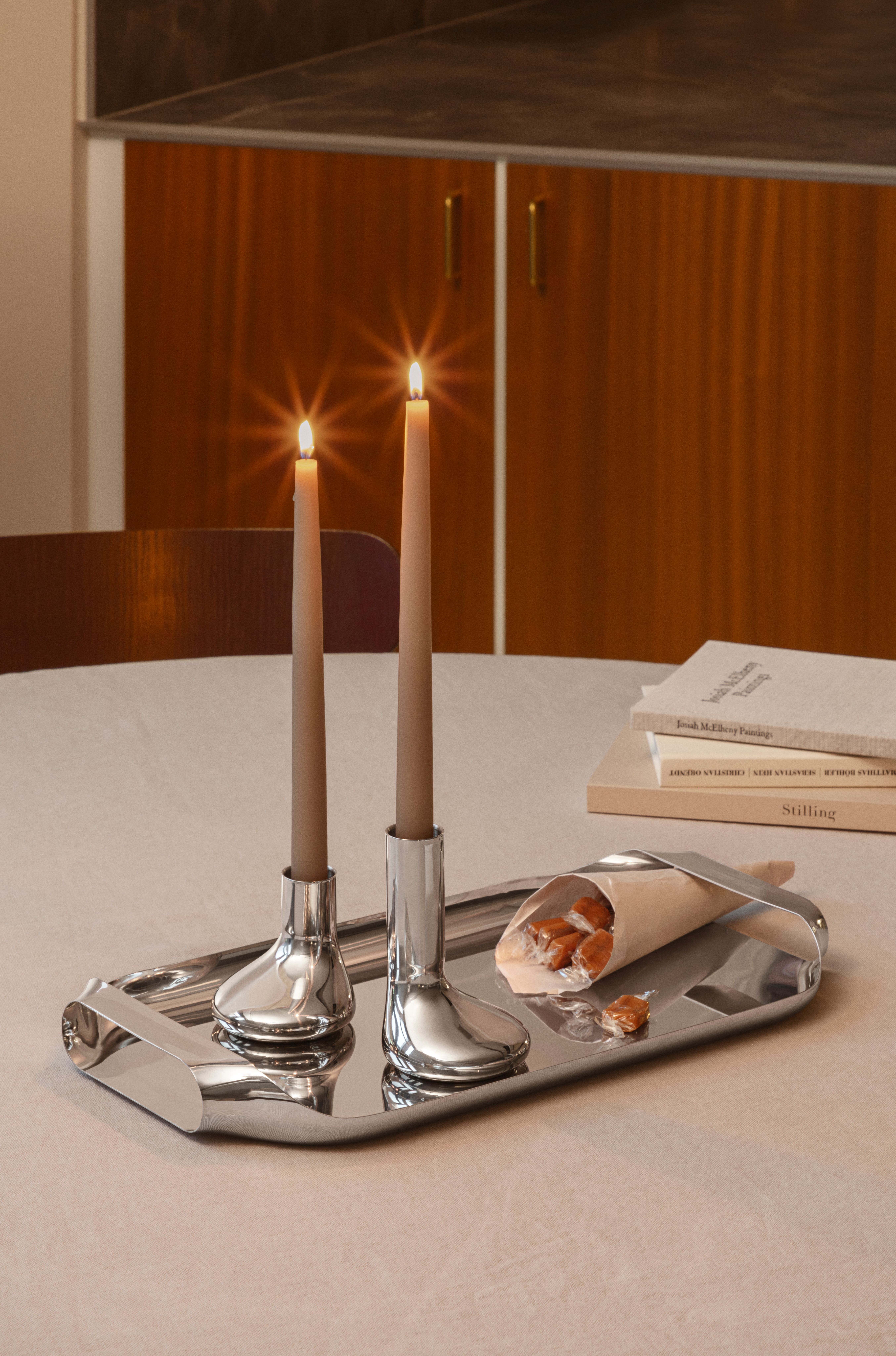 Two lit brown candles in silver holders on a tray with candies and stacked books.