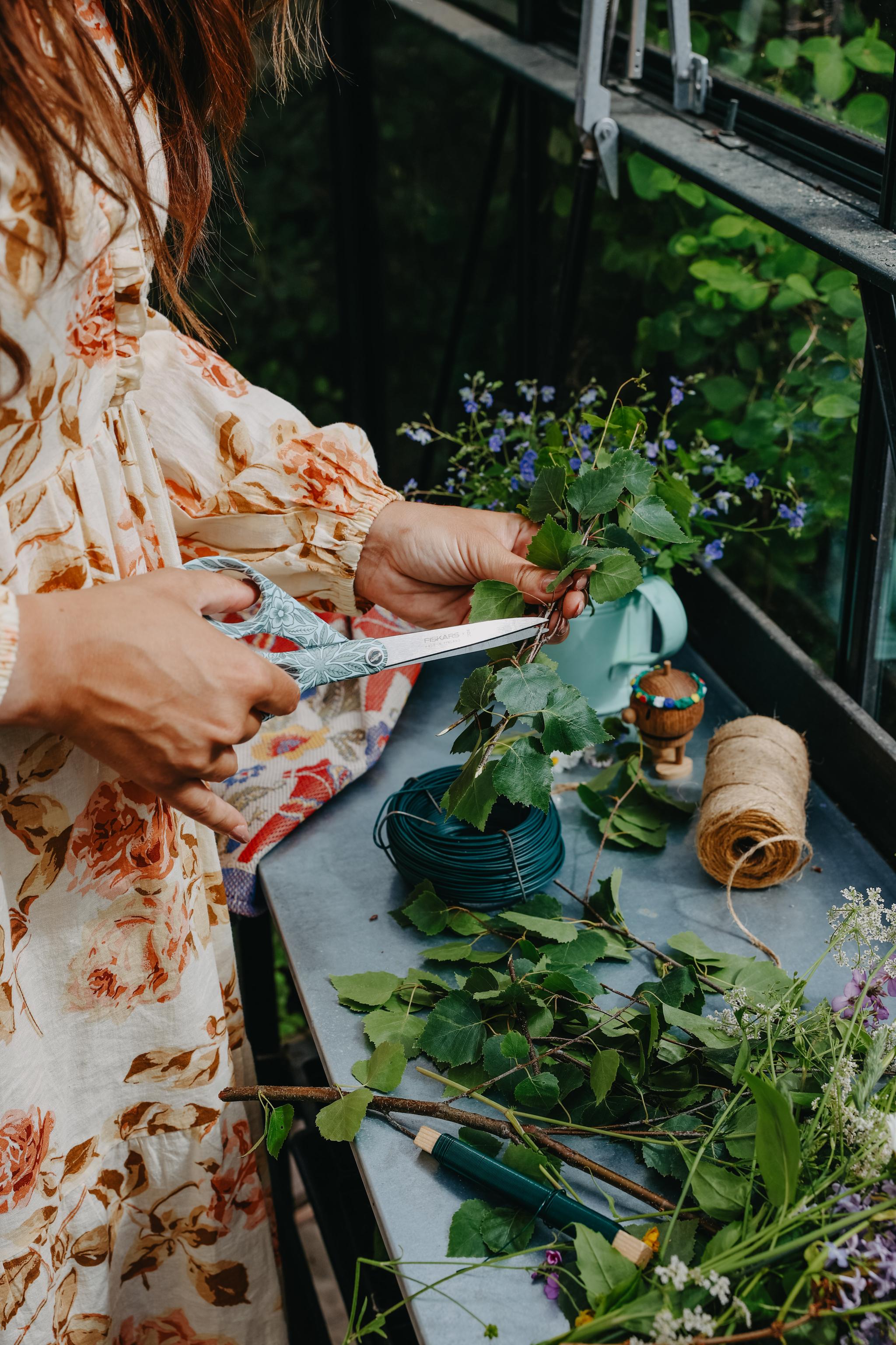 Person i blommig klänning binder en sommarkrans med gröna björklöv och blommor på ett bord i växthus.