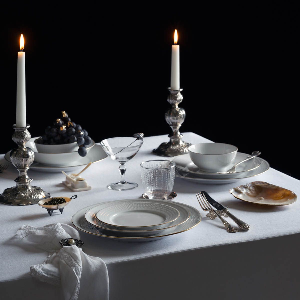 A dinner table set with two lit candles, fine china, silverware, grapes with gold leaf, a kintsugi glass, and caviar, on a white tablecloth against a dark background.