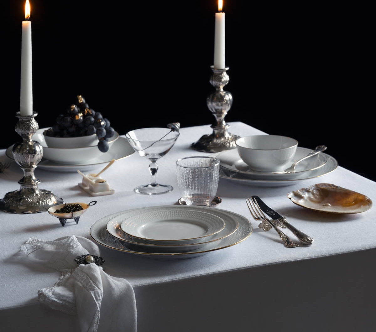 Et formelt dekket spisebord med tente lys i utsmykkede sølvstaker, elegante hvite tallerkener med gullkant, en skål med druer dekorert med bladgull, kaviar og et glass reparert med kintsugi, på en hvit duk mot en mørk bakgrunn.