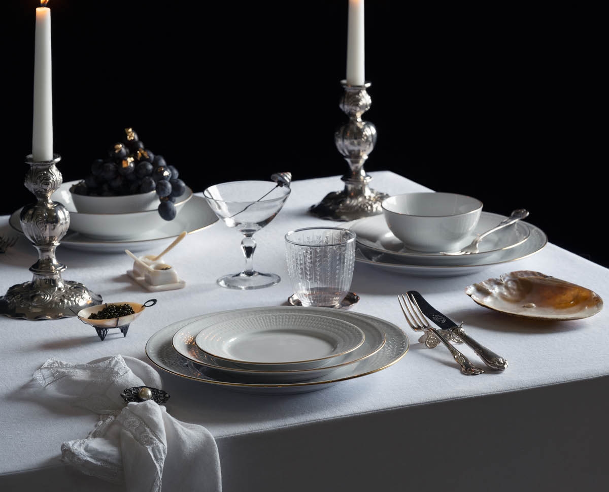 A dinner table set with two lit candles, fine china, silverware, grapes with gold leaf, a kintsugi glass, and caviar, on a white tablecloth against a dark background.