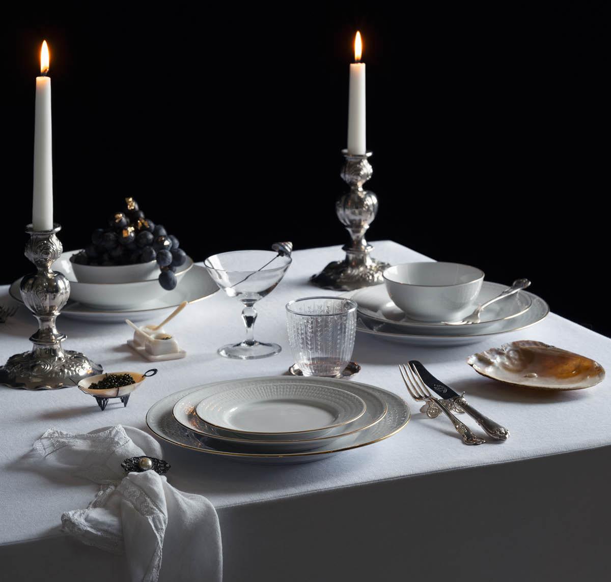 Et formelt dekket spisebord med tente lys i utsmykkede sølvstaker, elegante hvite tallerkener med gullkant, en skål med druer dekorert med bladgull, kaviar og et glass reparert med kintsugi, på en hvit duk mot en mørk bakgrunn.