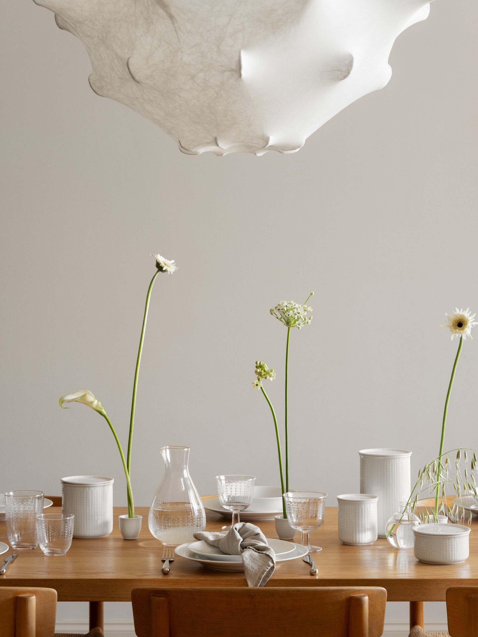 A modern dining table is set with white dishes, clear glassware, and delicate flowers, beneath a sculptural white light fixture.