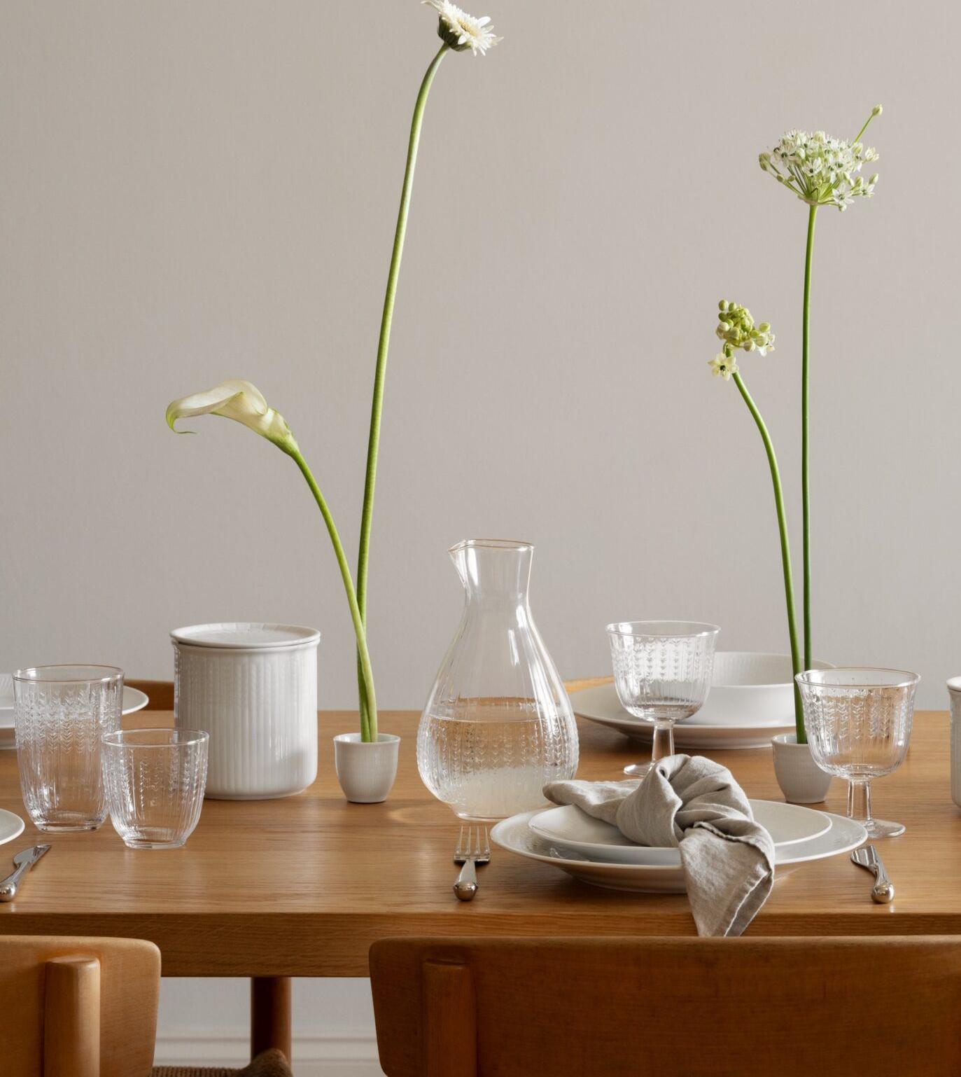 Et trebord er dekket med hvitt porselen, klare glass og fine hvite blomster i smale vaser, under en organisk hvit lampe.