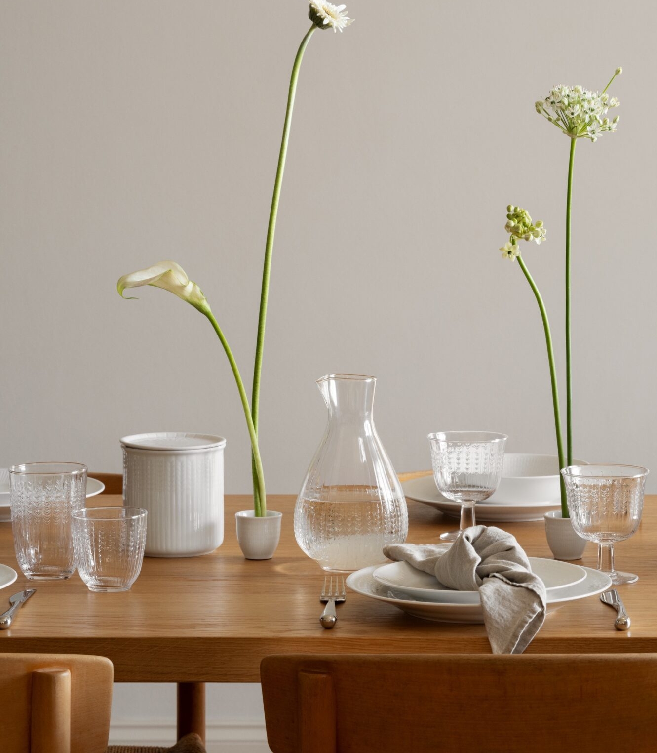 Et trebord er dekket med hvitt porselen, klare glass og fine hvite blomster i smale vaser, under en organisk hvit lampe.