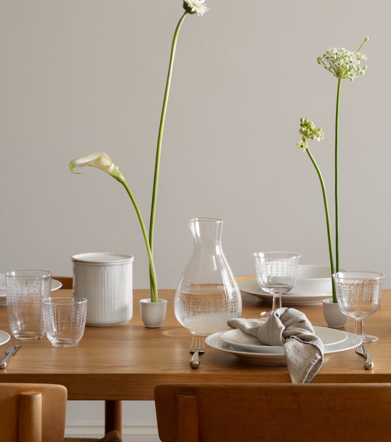Ett matbord i trä är dukat med vitt porslin, klara glas och fina vita blommor i smala vaser, under en organisk vit lampa.