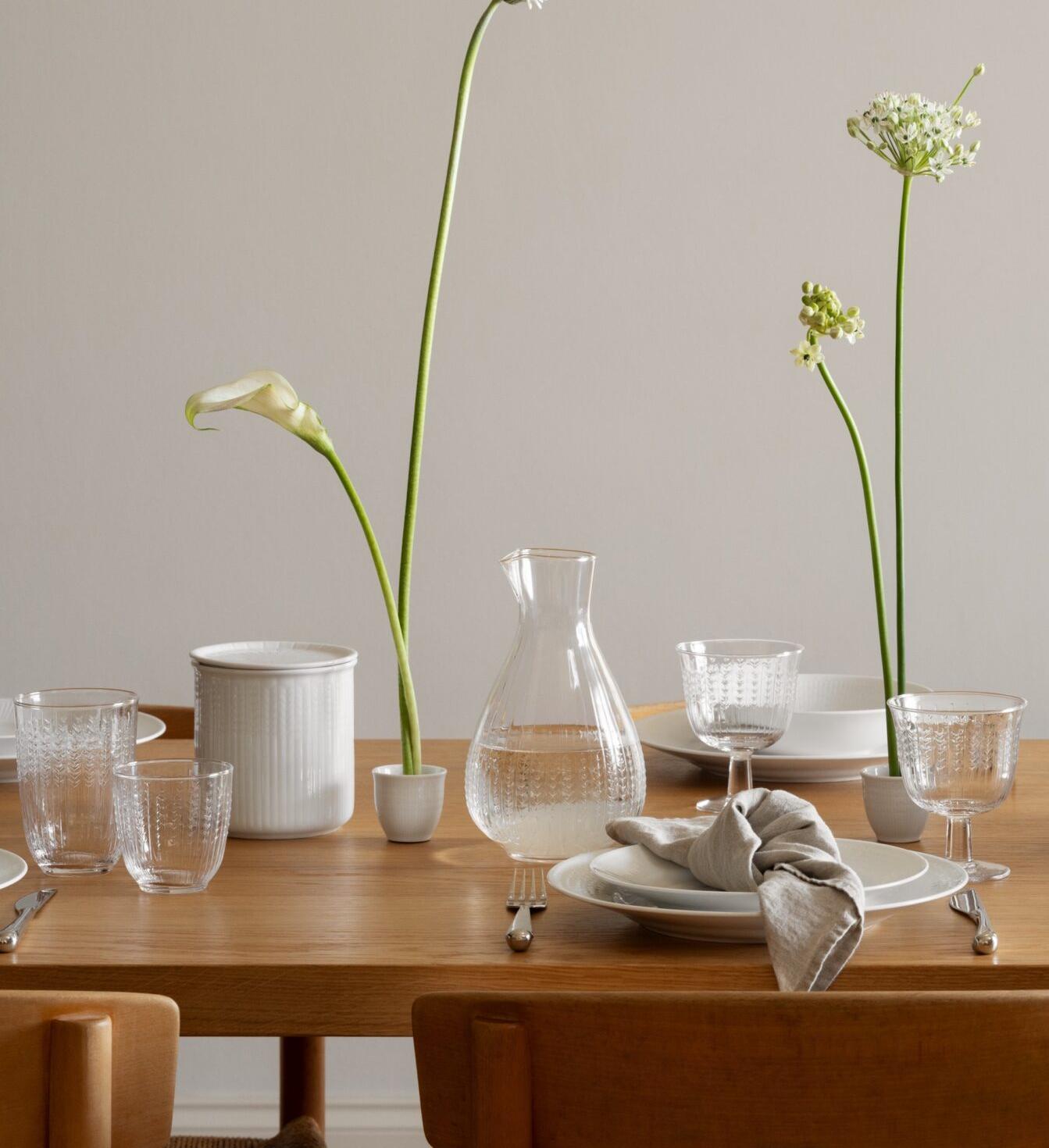 A modern dining table is set with white dishes, clear glassware, and delicate flowers, beneath a sculptural white light fixture.