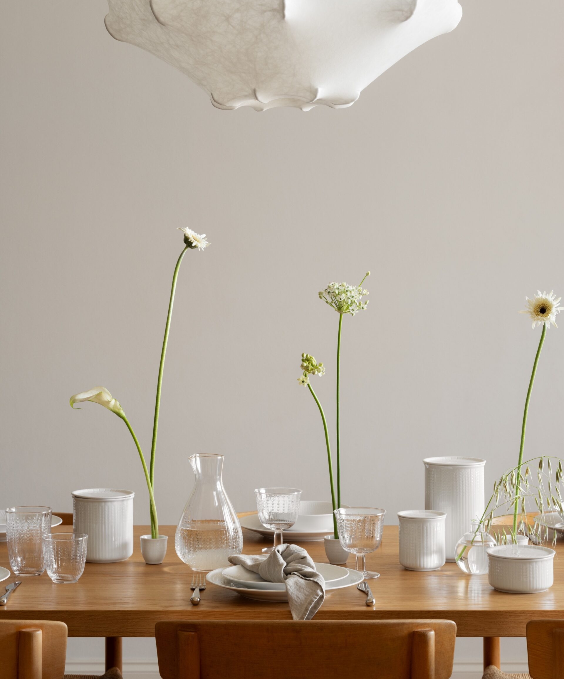 Elegant dukning på träbord med vitt porslin, glas och höga vita blommor i en ljus matsal.
