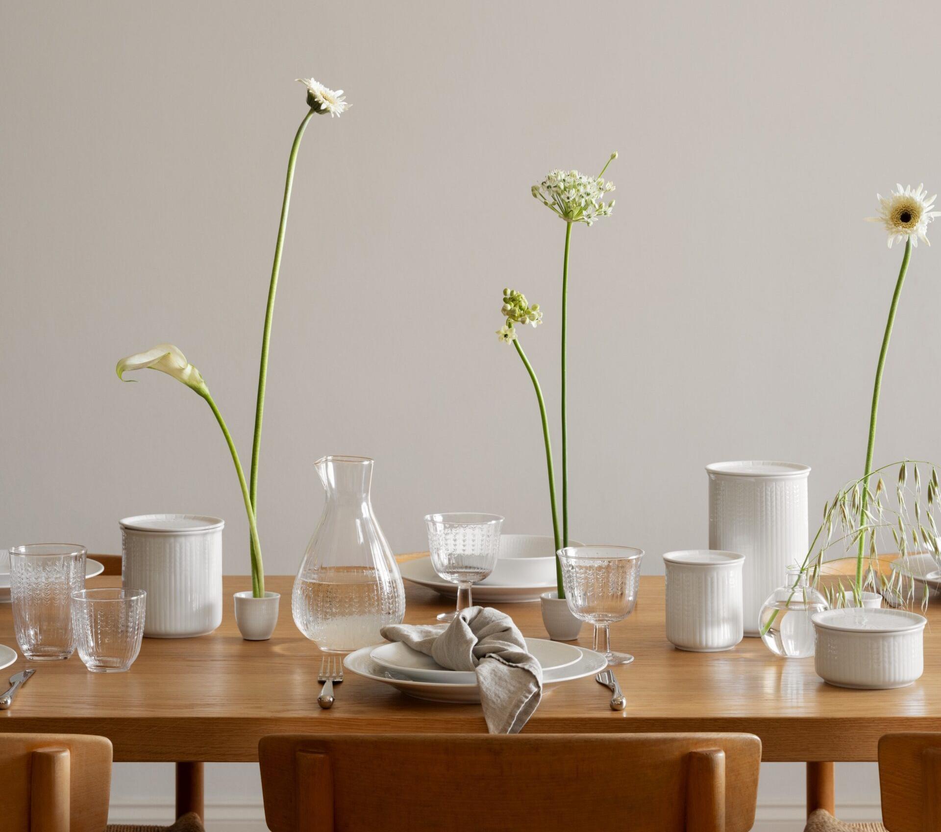 Elegant dukning på träbord med vitt porslin, glas och höga vita blommor i en ljus matsal.