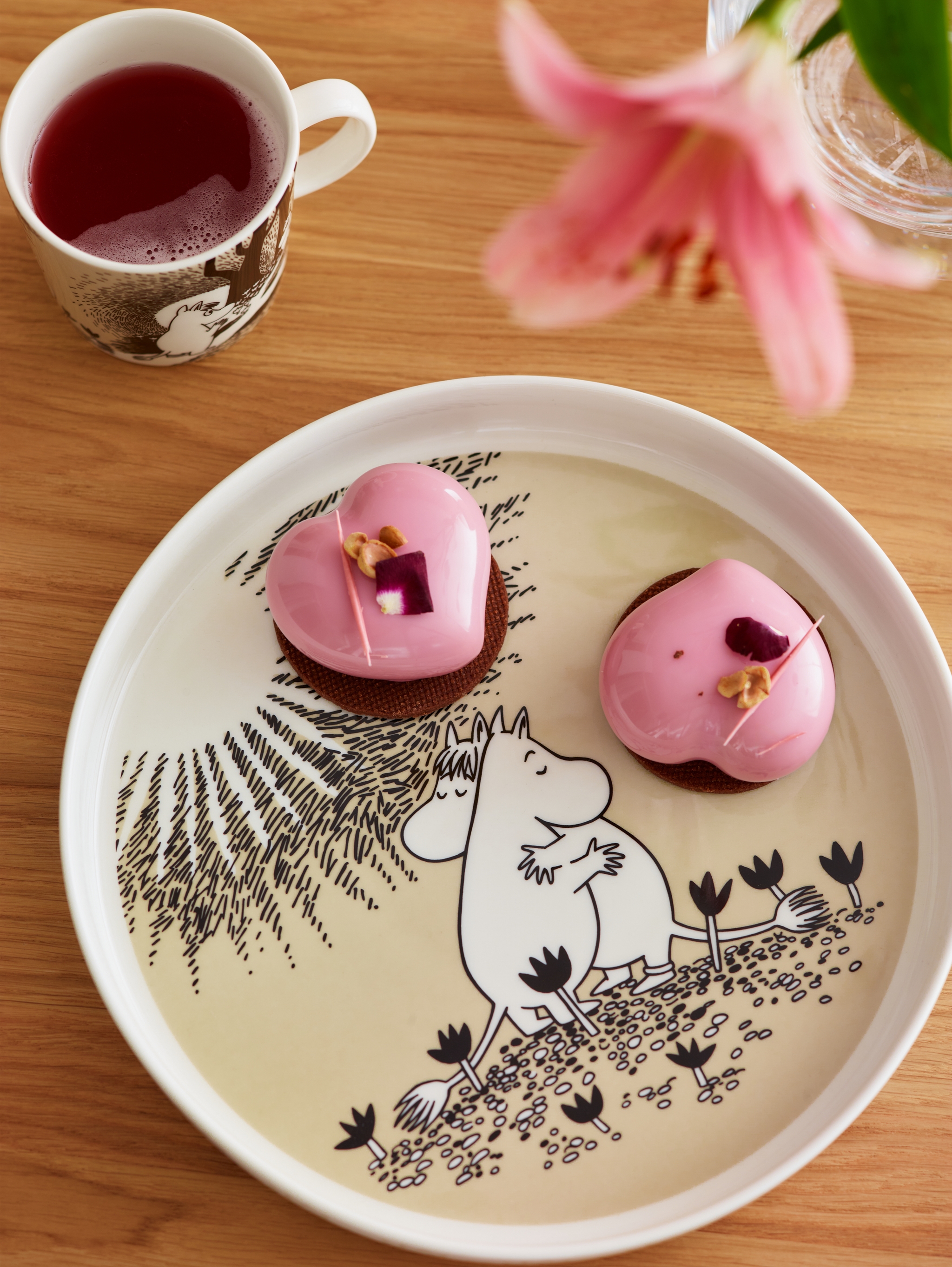 Rosa hjärtformade bakelser på ett Mumin-fat med en kopp rött te och en rosa blomma på ett träbord.