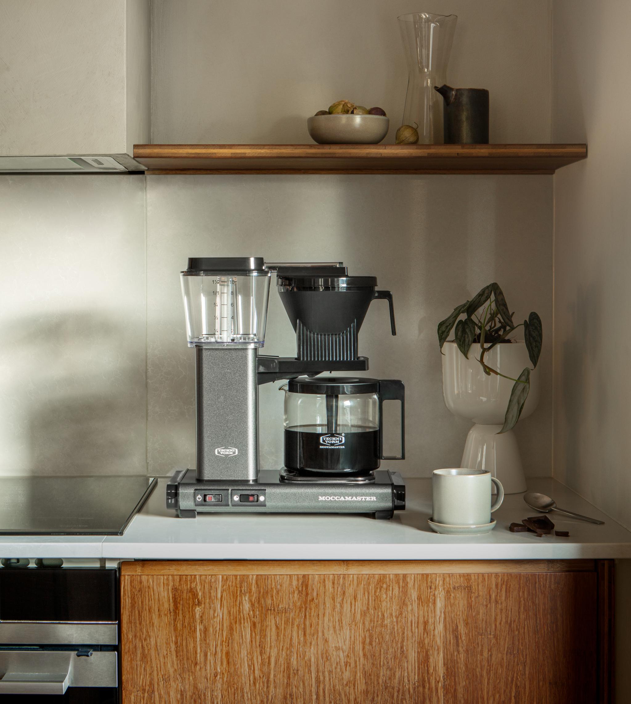 Modern kitchen counter with a grey Moccamaster coffee maker, a white mug, and a plant, below a wooden shelf holding fruit and glassware.