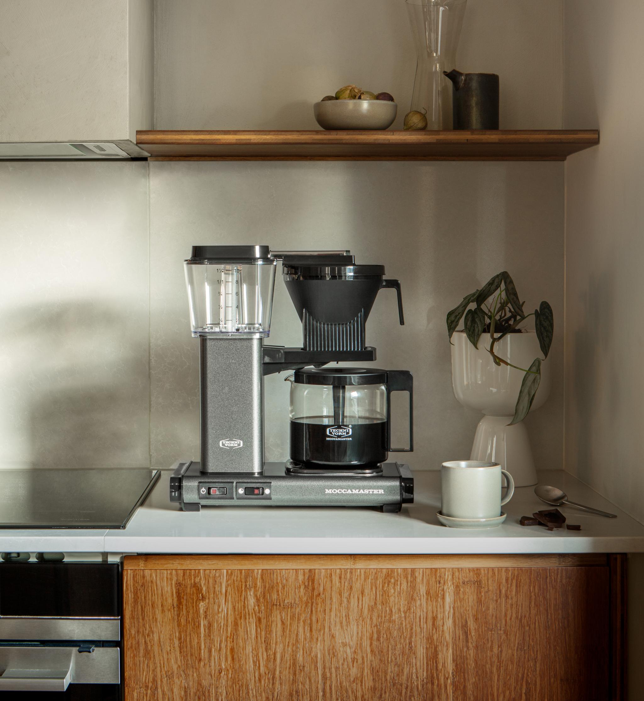 Moccamaster kaffetrakter på en stilren kjøkkenbenk med kopp, planter og fruktbolle i bakgrunnen.