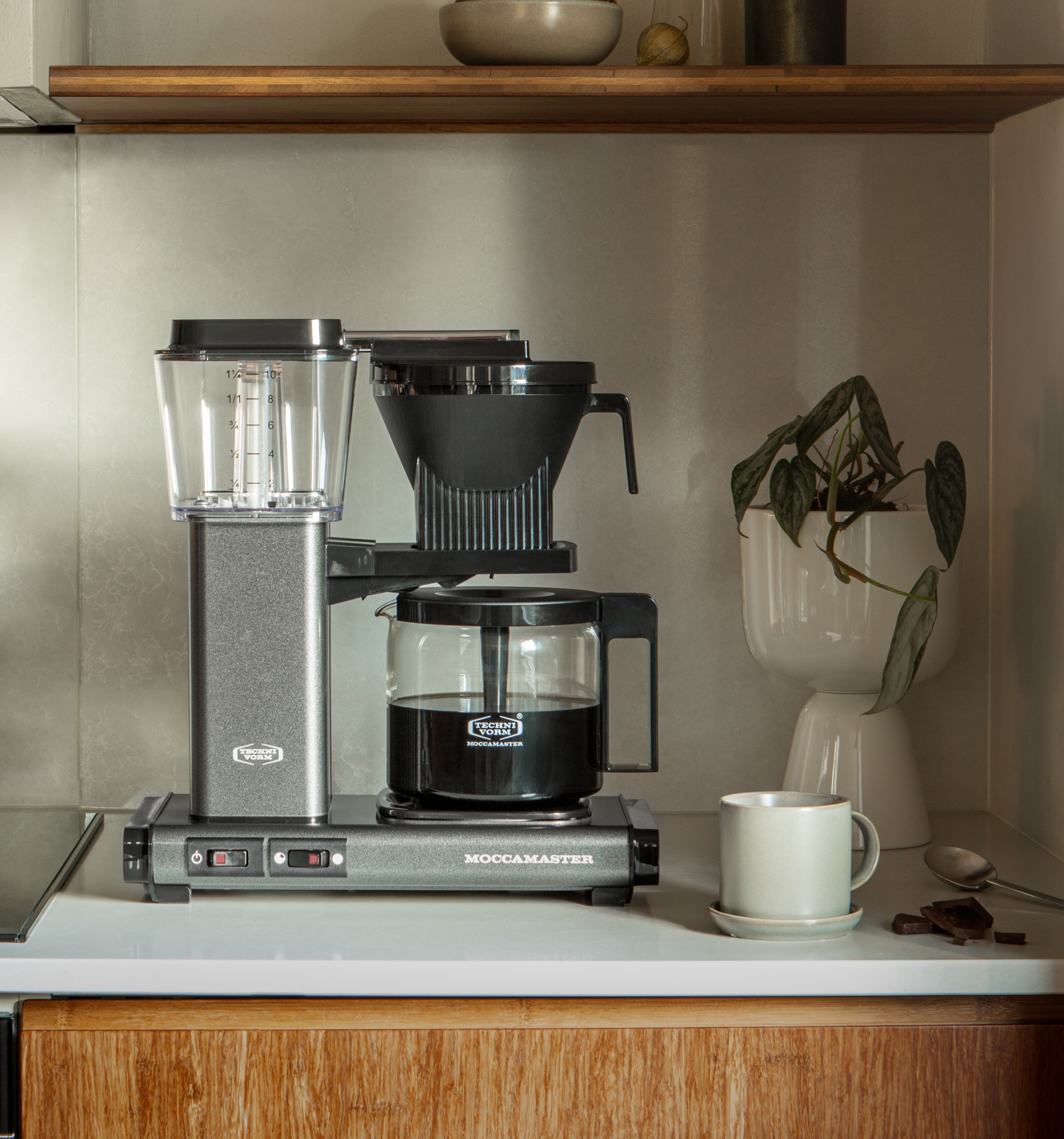 Stilren kjøkkenbenk med Moccamaster kaffetrakter, kaffekopp og grønne planter i et moderne, nordisk interiør.