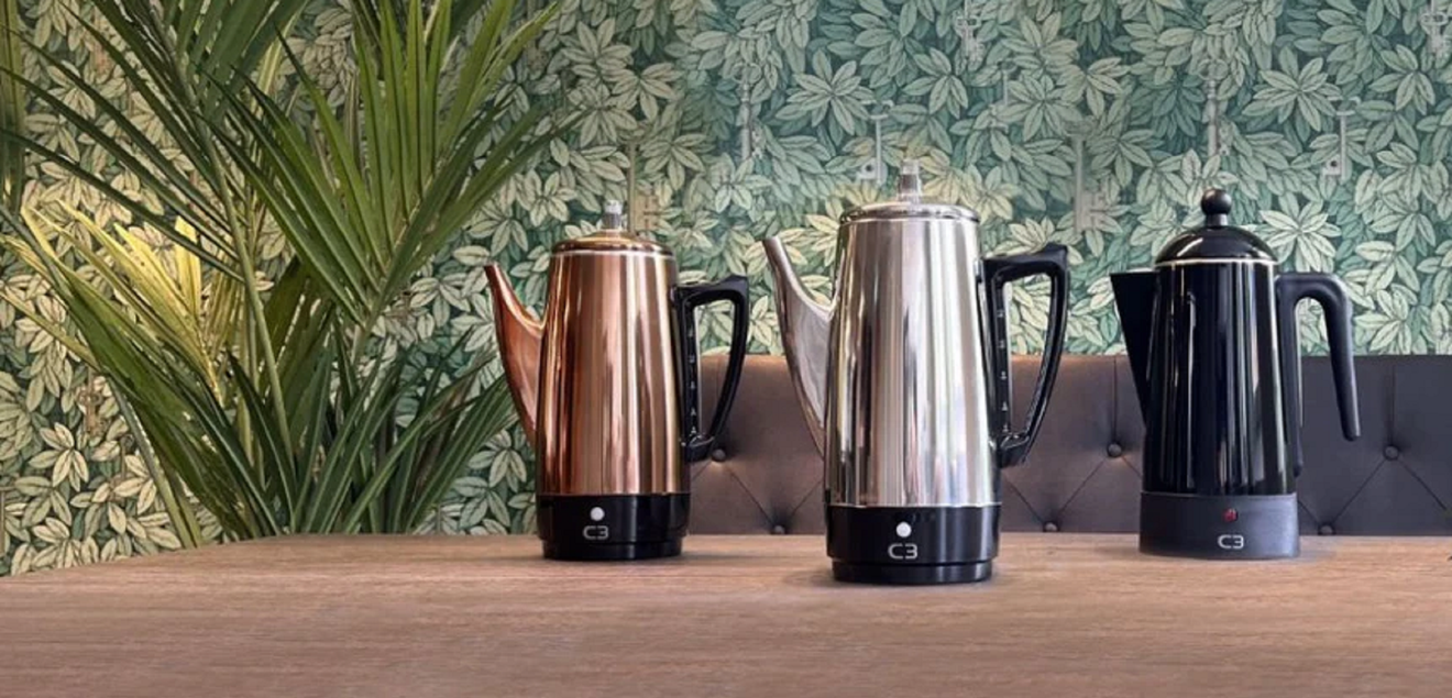 Three electric percolator coffee makers in copper, silver, and black on a wooden table.