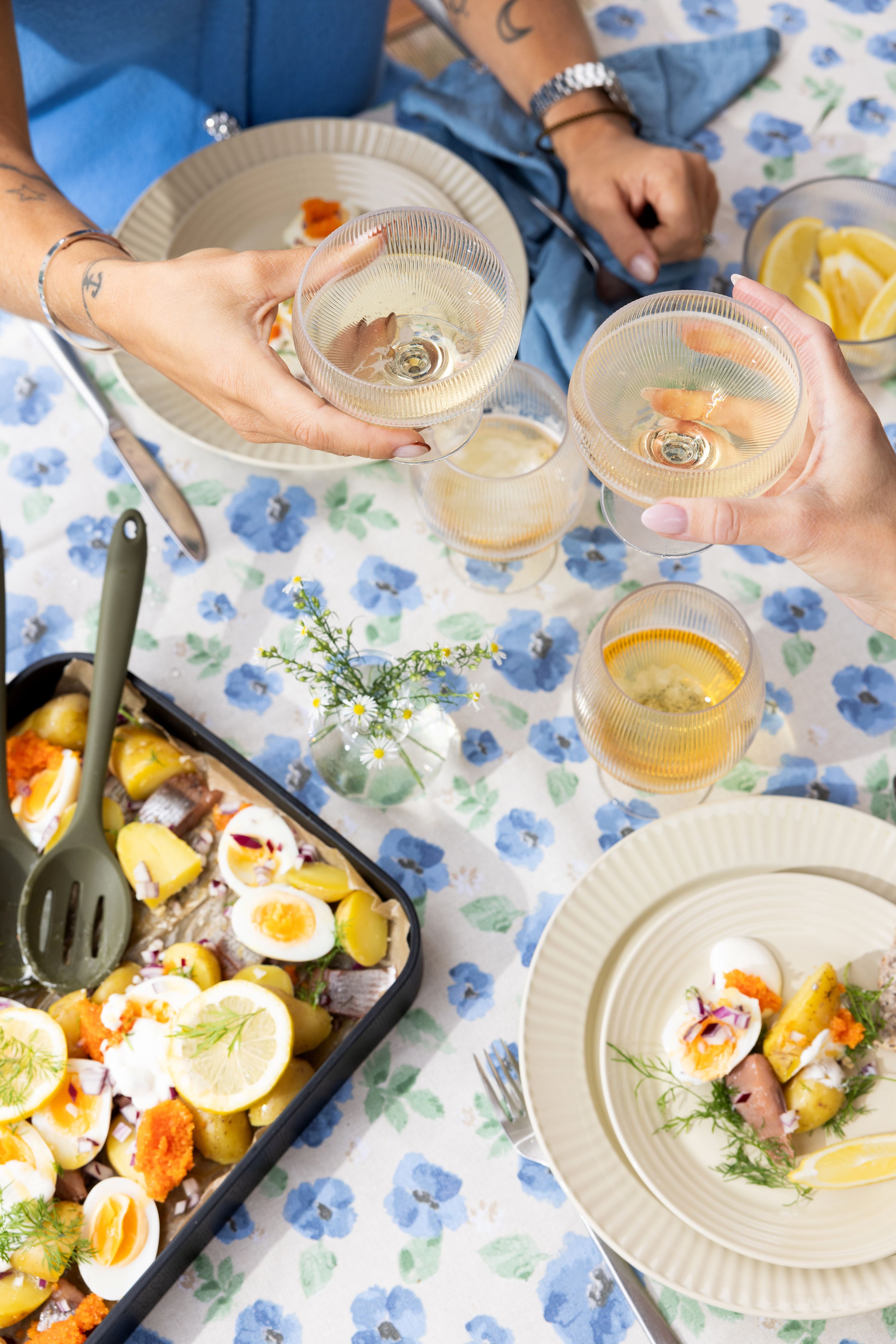 Två personer skålar med vinglas över en blå blommig duk med en måltid bestående av färskpotatis, hårdkokta ägg, inlagd sill och rom