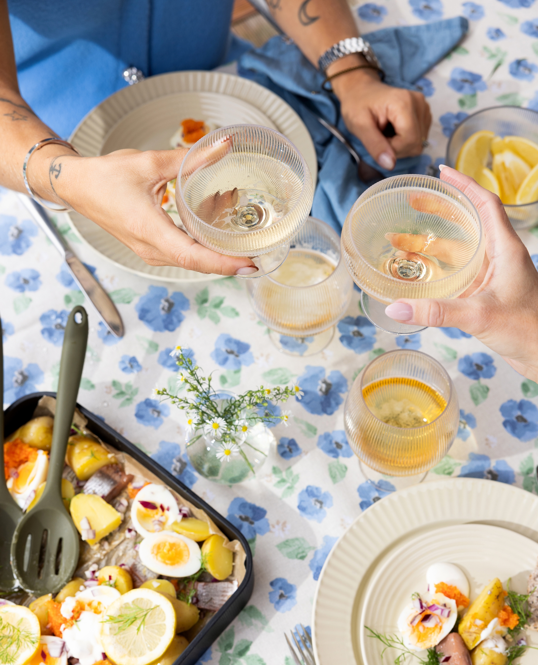 Två personer skålar med vinglas över en blå blommig duk med en måltid bestående av färskpotatis, hårdkokta ägg, inlagd sill och rom