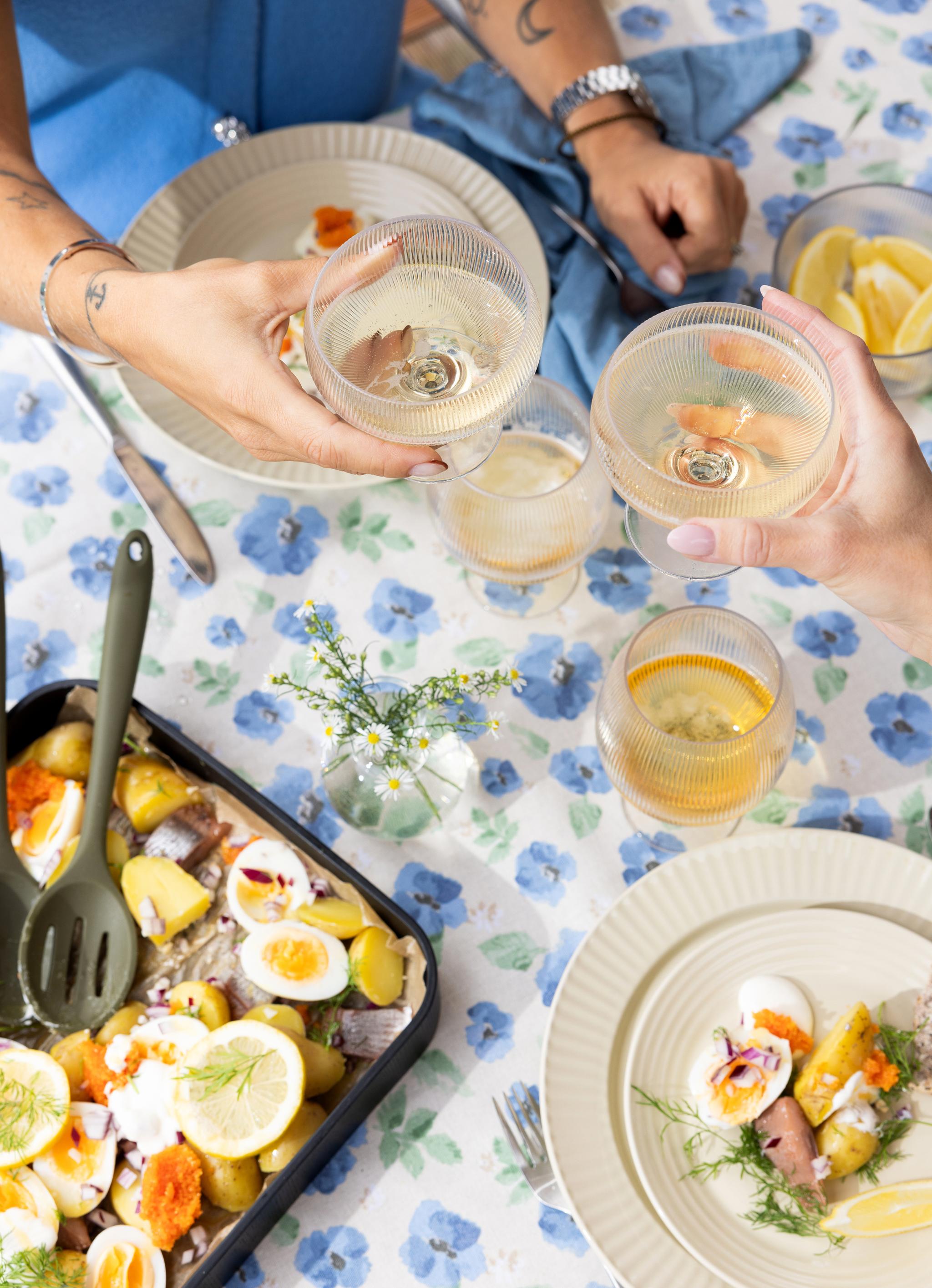 Två personer skålar med vinglas över en blå blommig duk med en måltid bestående av färskpotatis, hårdkokta ägg, inlagd sill och rom
