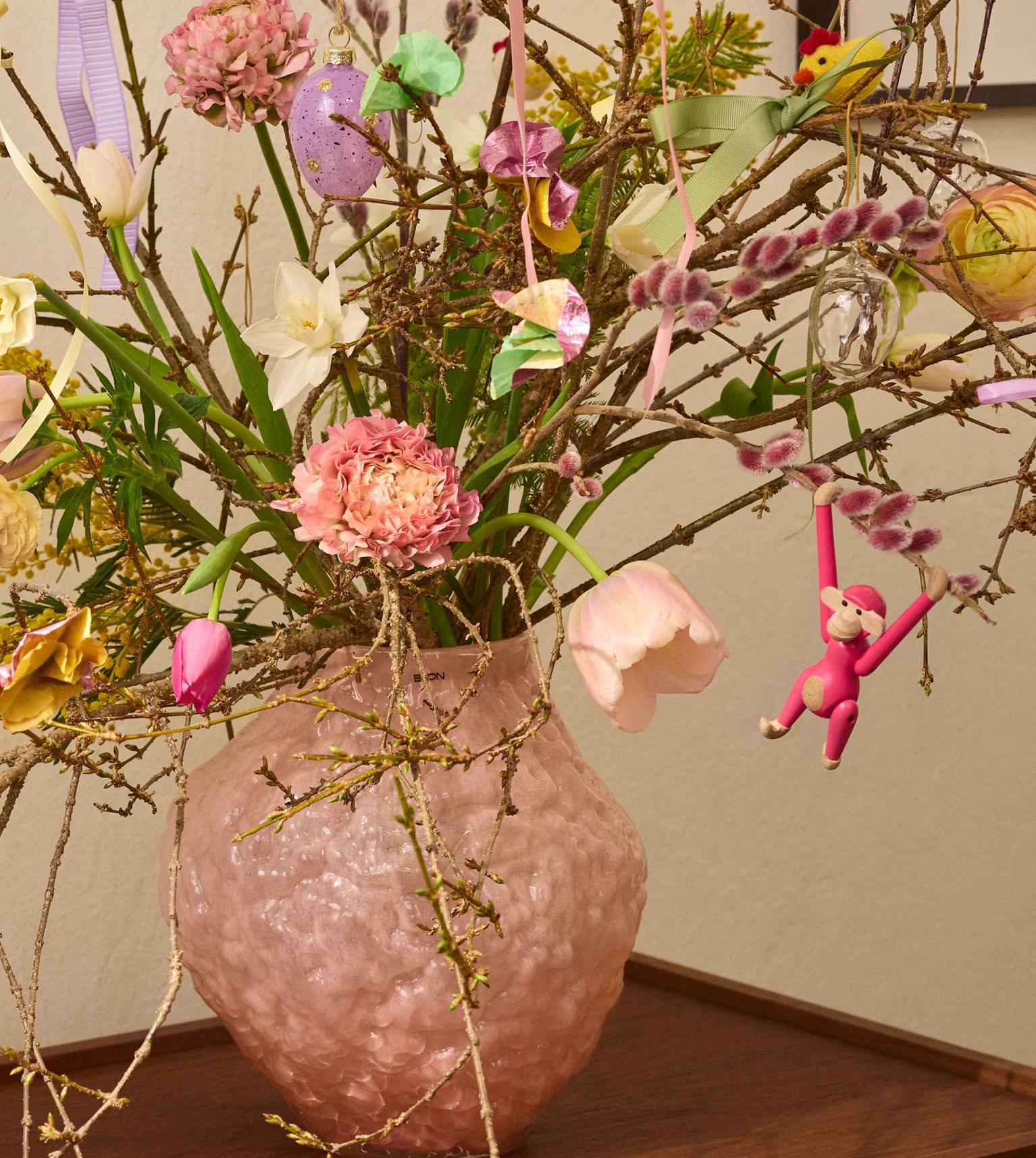 A pink vase on a wooden table holds branches and spring flowers decorated with colorful Easter eggs, ribbons, and small animal ornaments.
