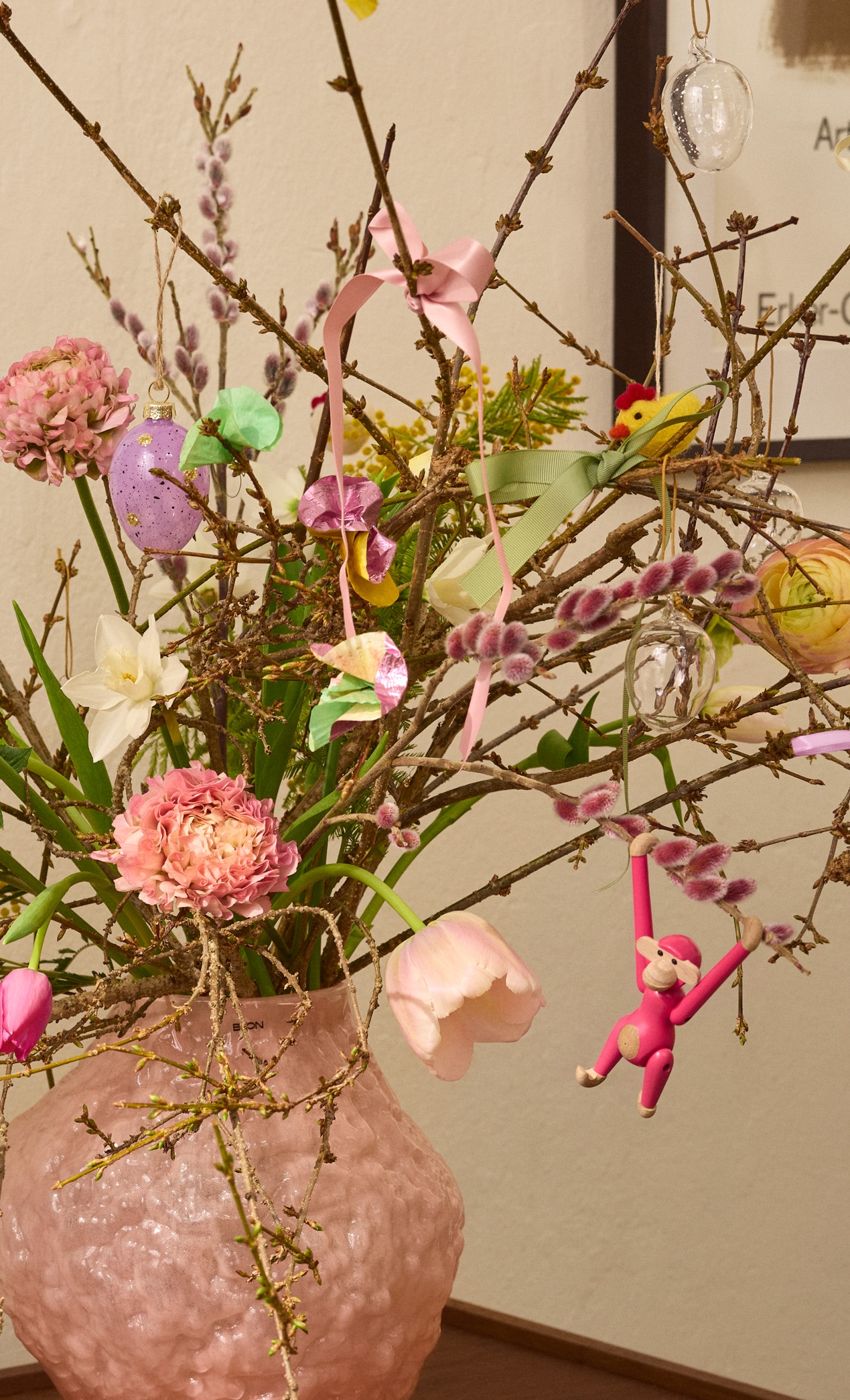 A pink textured vase holds spring branches decorated with Easter eggs, bunnies, ribbons, and fresh flowers on a wooden table.