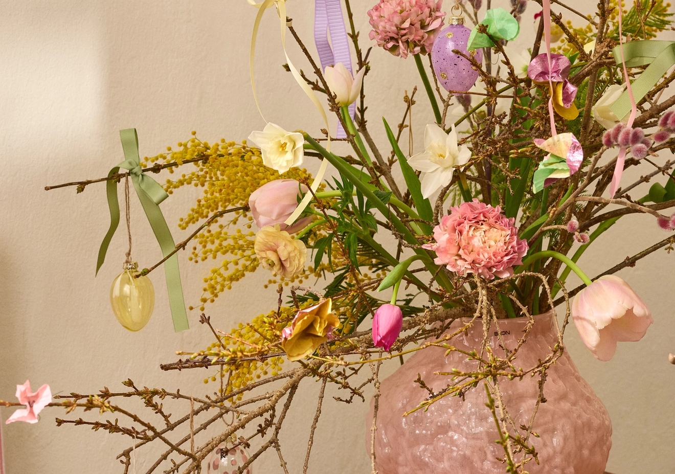 En rosa texturerad vas håller grenar dekorerade med färgglada påskägg, kaniner, band och färska blommor inklusive mimosa, påskliljor och tulpaner, på ett träbord.