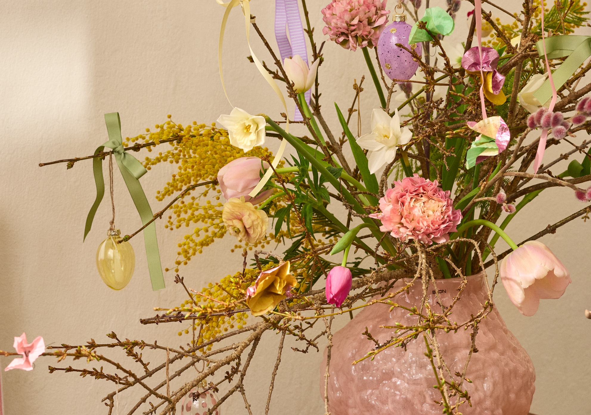 En rosa texturerad vas håller grenar dekorerade med färgglada påskägg, kaniner, band och färska blommor inklusive mimosa, påskliljor och tulpaner, på ett träbord.