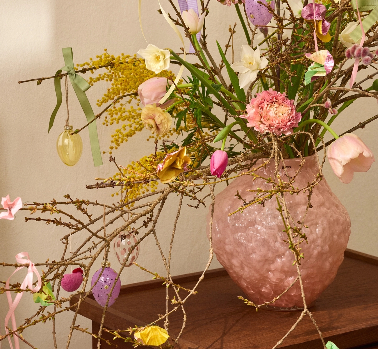 En rosa texturerad vas håller grenar dekorerade med färgglada påskägg, kaniner, band och färska blommor inklusive mimosa, påskliljor och tulpaner, på ett träbord.