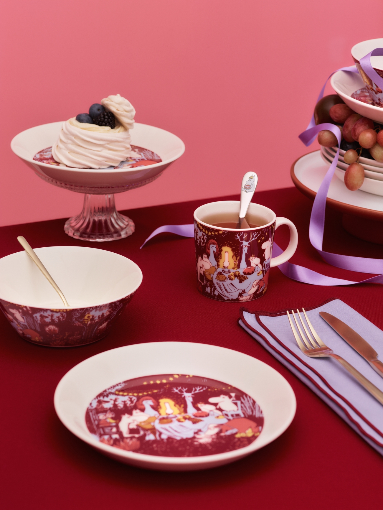 A festive table setting with white dishes featuring dark red illustrations, a meringue dessert, and a mug of tea, against a red tablecloth and pink background.