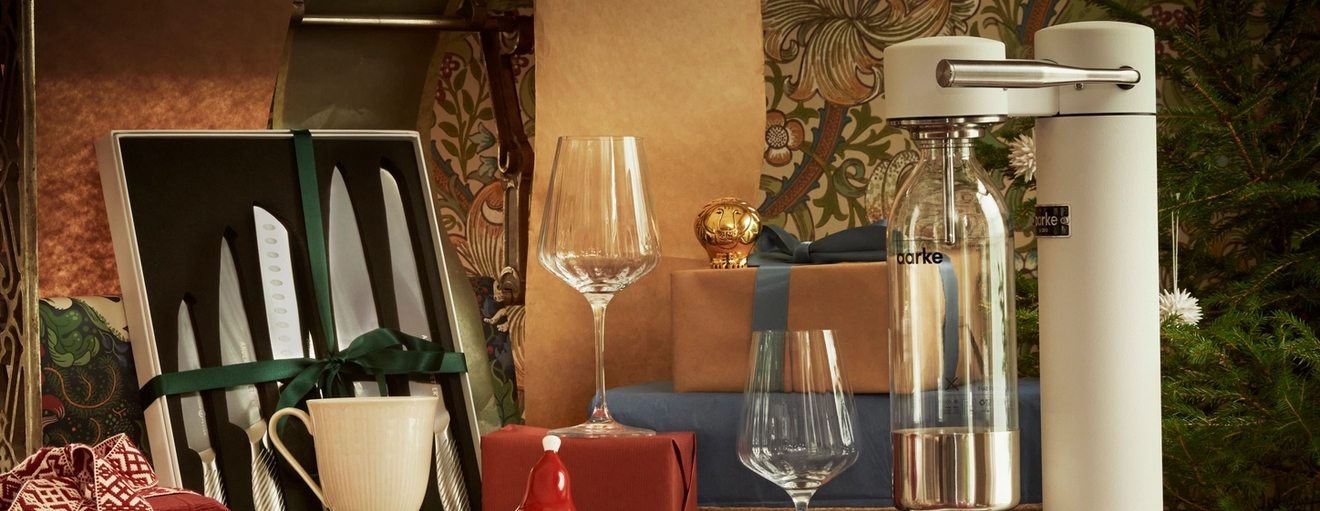 Festlig utstilling av kjøkkenutstyr og gaver, med et knivsett, vinglass, innpakkede esker og en brusmaskin ved siden av et juletre.