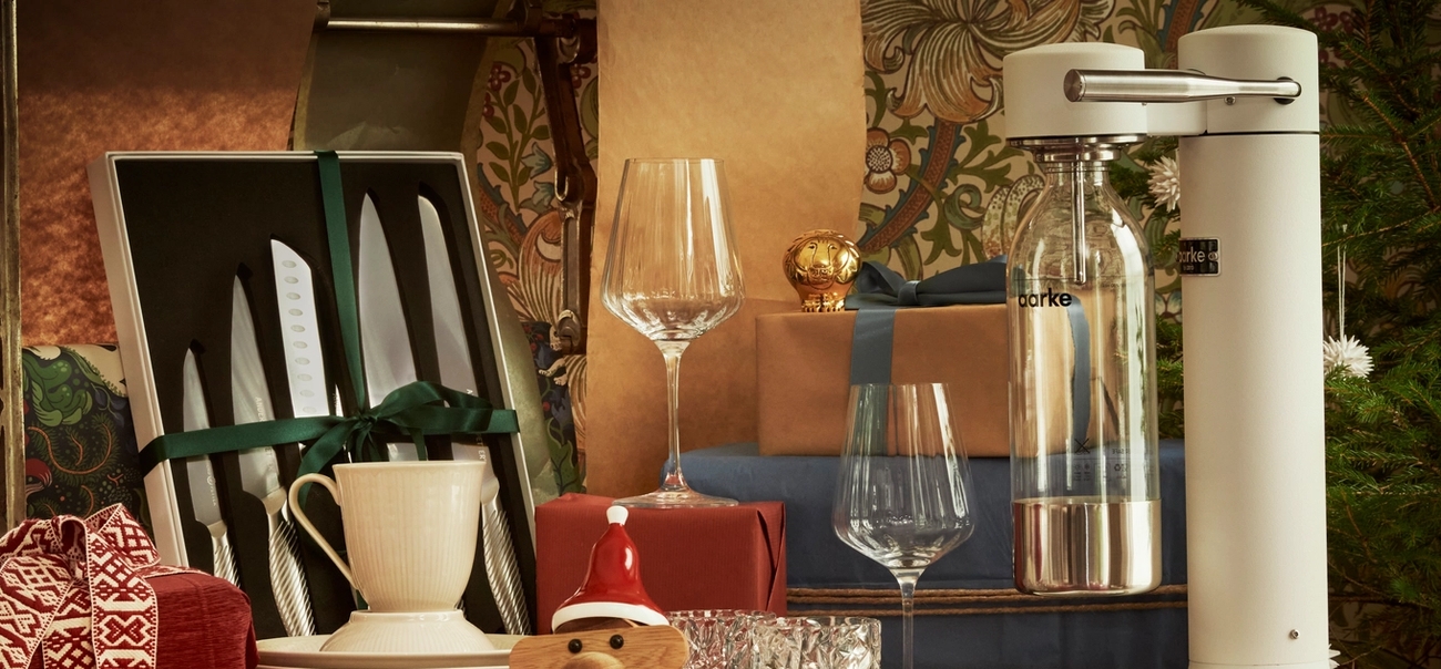 Festlig utstilling av kjøkkenutstyr og gaver, med et knivsett, vinglass, innpakkede esker og en brusmaskin ved siden av et juletre.