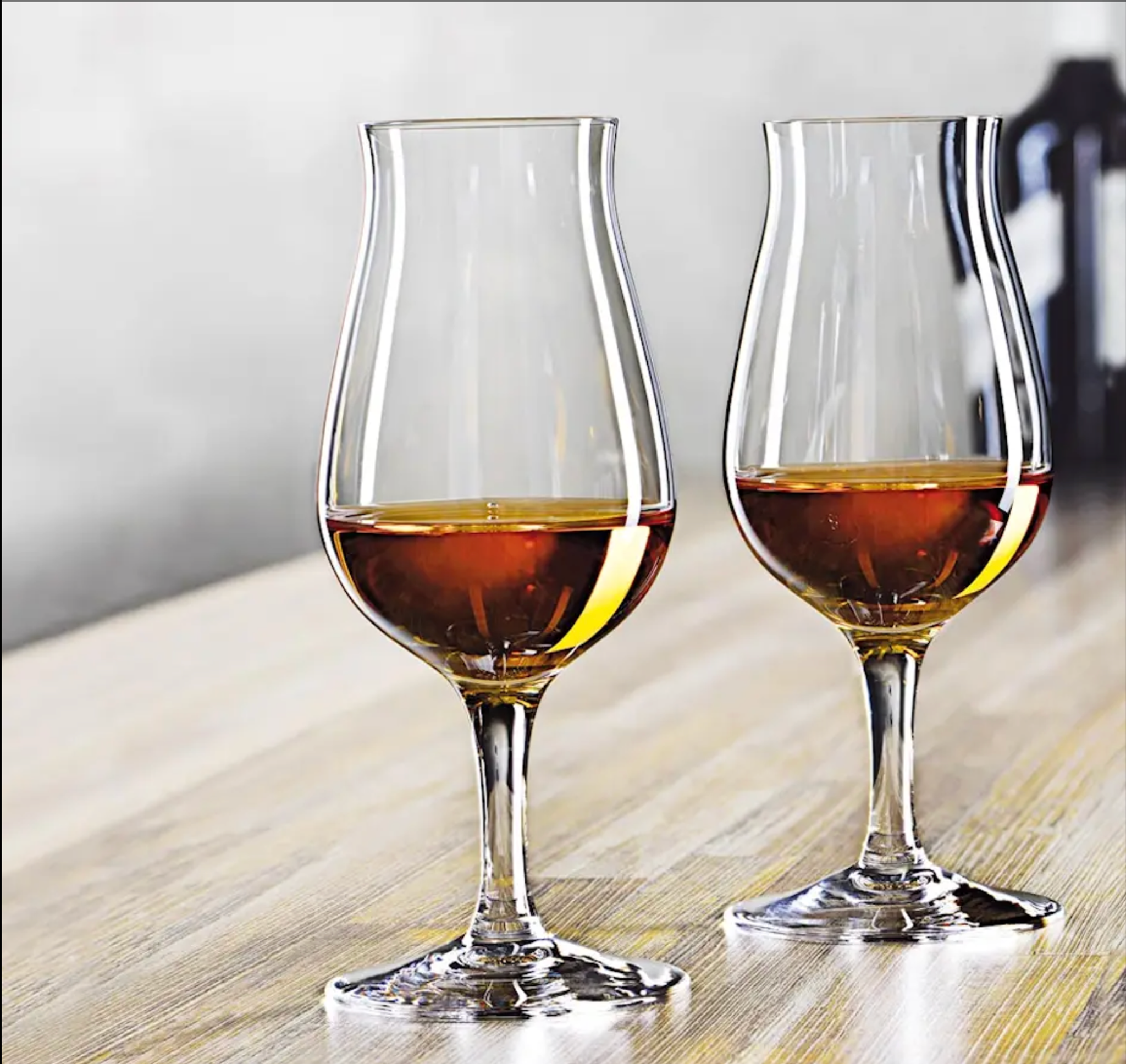 Två whiskyglas med gyllenbrun whiskey i sig, placerade på ett ljust träbord med en suddig bakgrund.