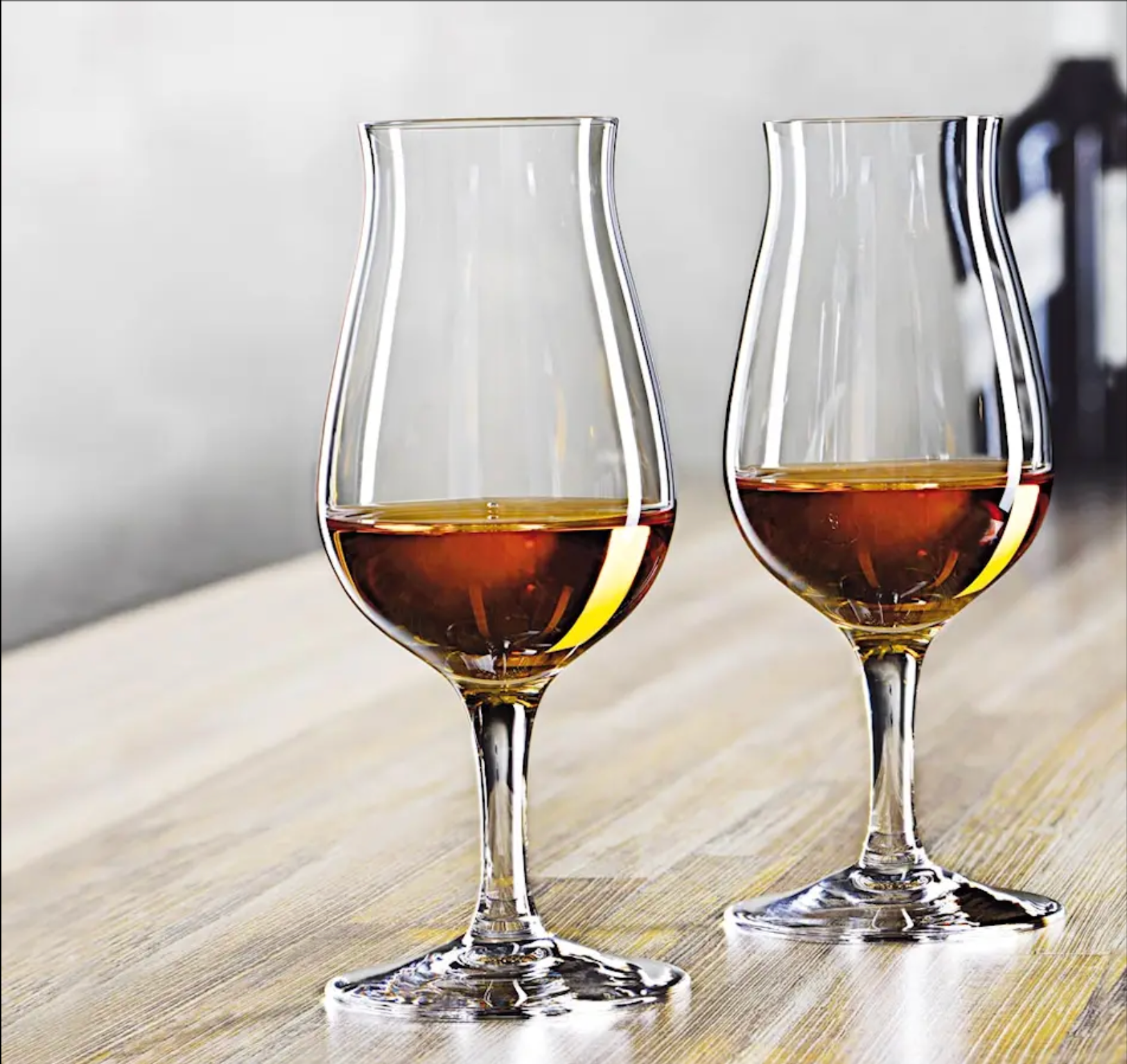 Två whiskyglas med gyllenbrun whiskey i sig, placerade på ett ljust träbord med en suddig bakgrund.