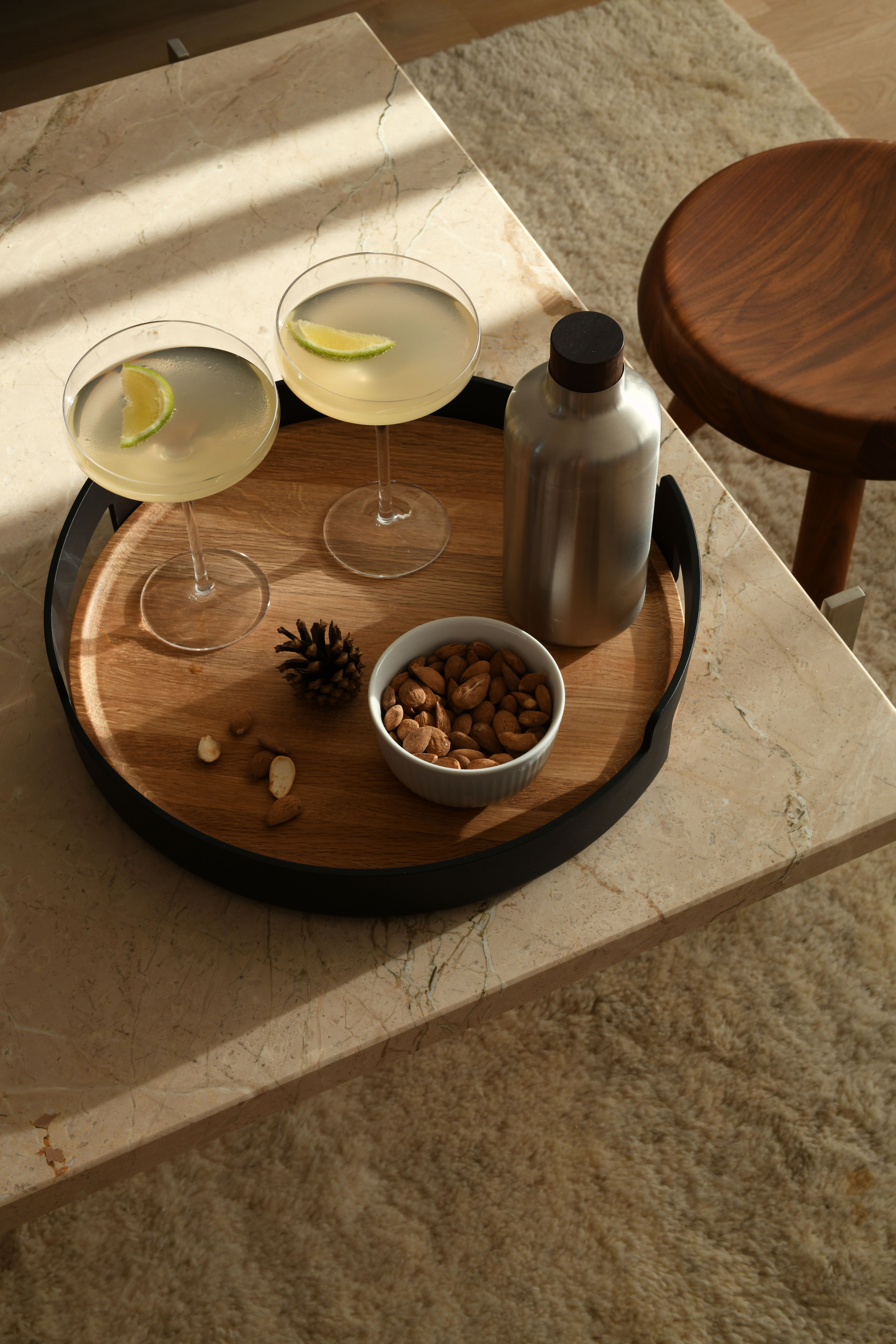 Kaksi limecocktailia, kulho manteleita ja cocktail-ravistin puisella tarjottimella marmorisella sohvapöydällä.