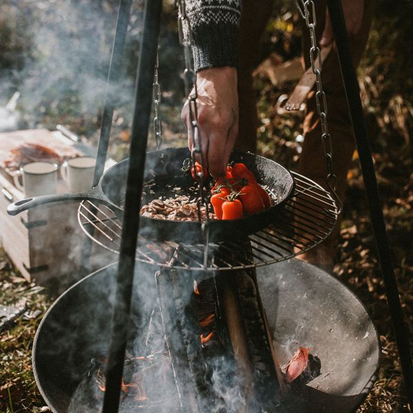 Henkilö valmistaa ruokaa ja grillaa tomaatteja valurautapannulla nuotion yllä ulkona.