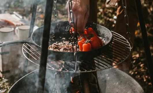 Henkilö valmistaa ruokaa ja grillaa tomaatteja valurautapannulla nuotion yllä ulkona.