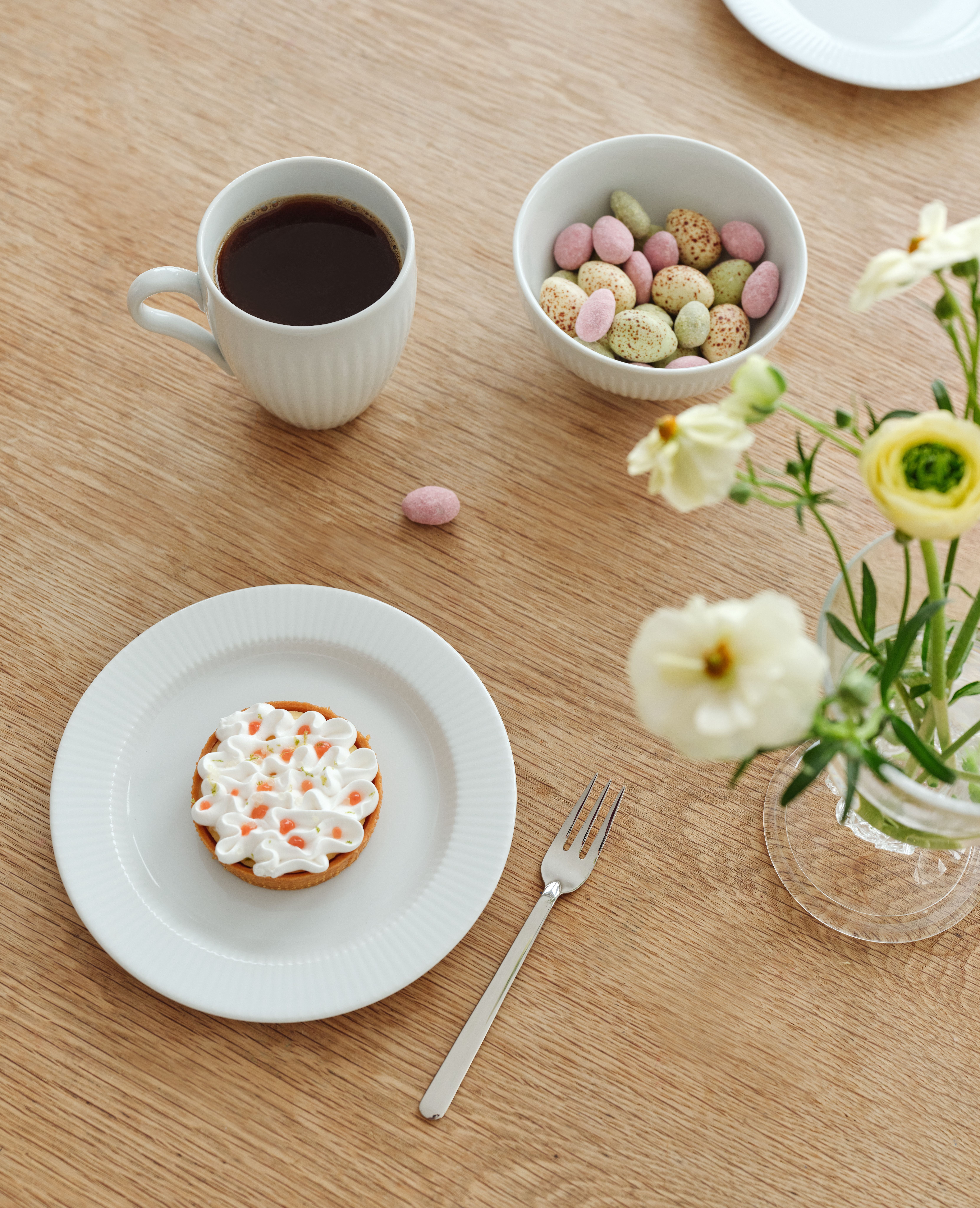 Vaalealla puupöydällä on muki kahvia, kulho pastellivärisiä munanmuotoisia karkkeja, marenkitorttu, haarukka ja maljakko, jossa on valkoisia ja keltaisia ​​kukkia.