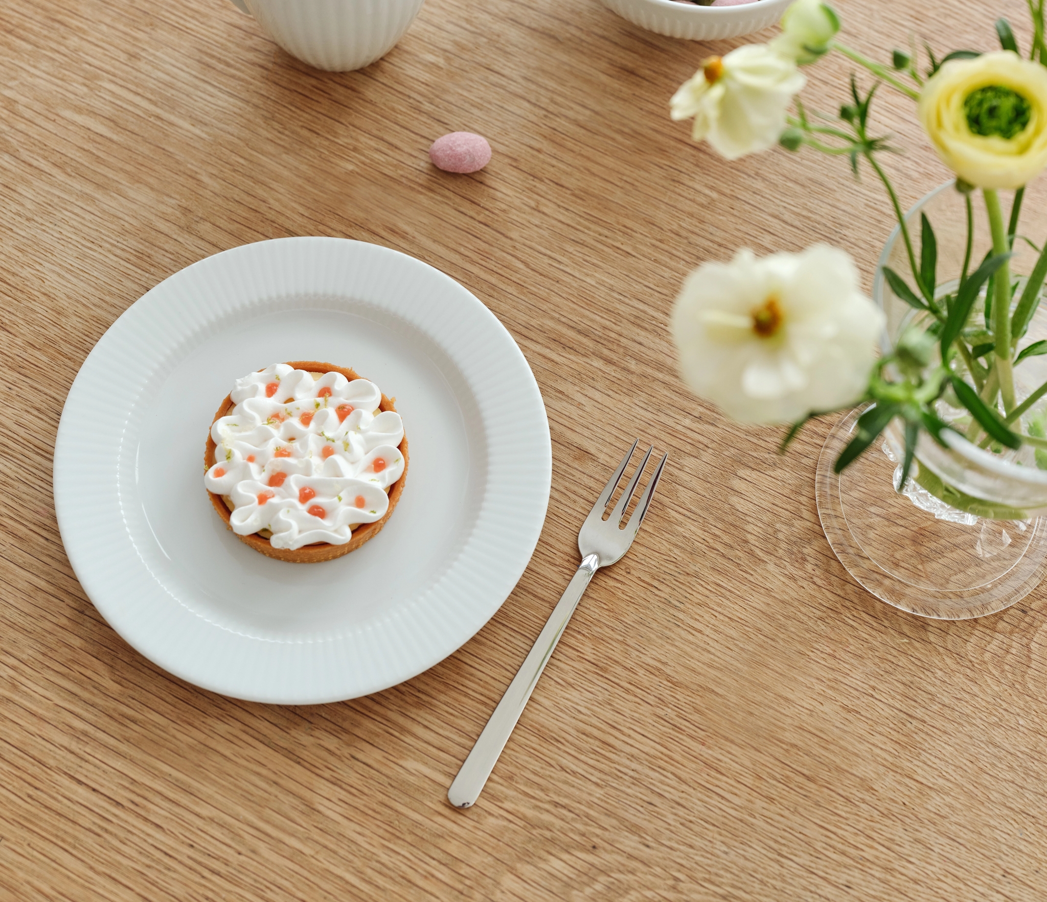 Vaalealla puupöydällä on muki kahvia, kulho pastellivärisiä munanmuotoisia karkkeja, marenkitorttu, haarukka ja maljakko, jossa on valkoisia ja keltaisia kukkia.