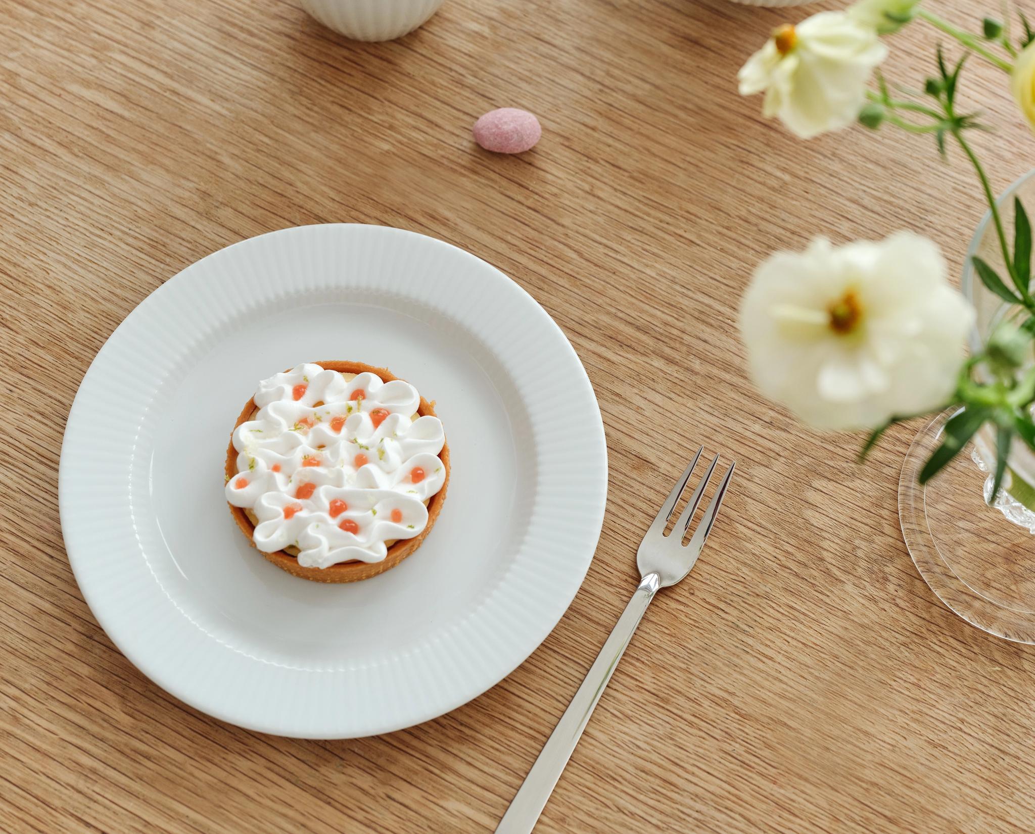 Vaalealla puupöydällä on muki kahvia, kulho pastellivärisiä munanmuotoisia karkkeja, marenkitorttu, haarukka ja maljakko, jossa on valkoisia ja keltaisia kukkia.