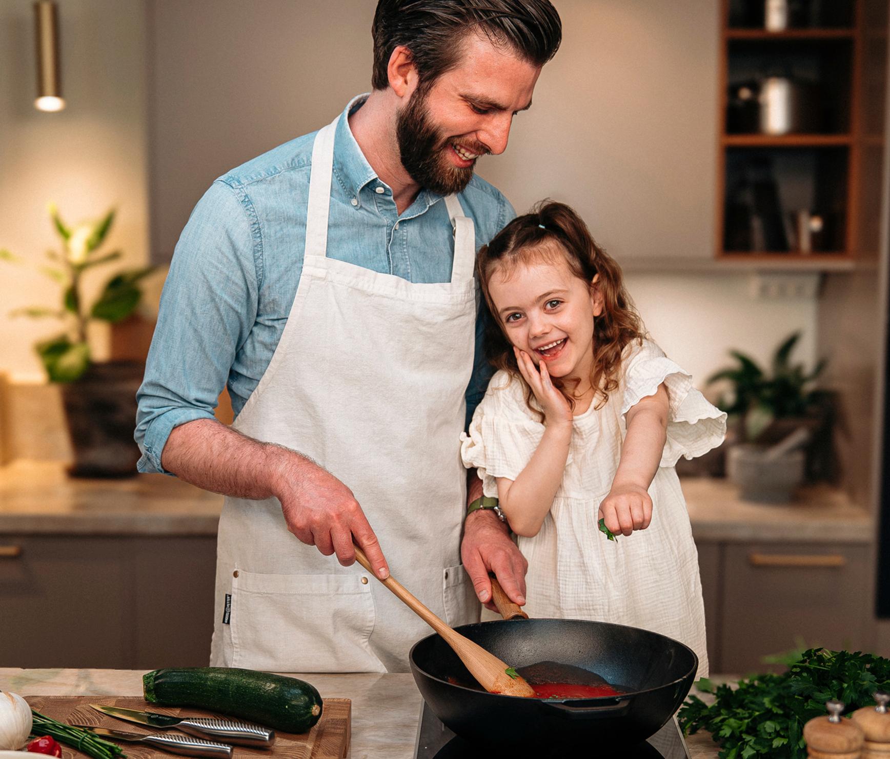 Leende pappa och dotter lagar mat tillsammans vid spisen i ett modernt kök, med Cervera-loggan ovanför.