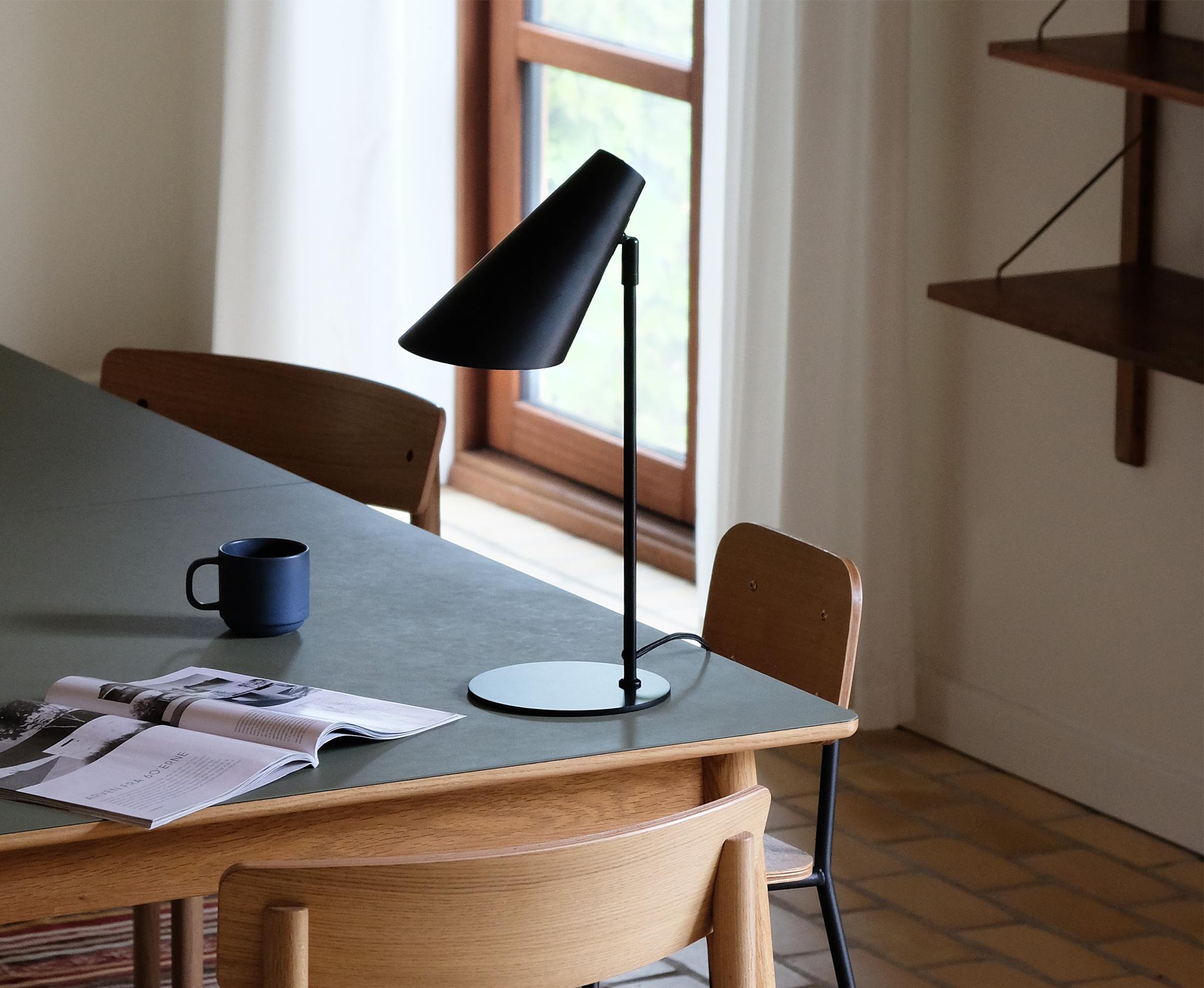 Ljust matrum med trästolar, grönt bord, svart bordslampa och ett öppet magasin.