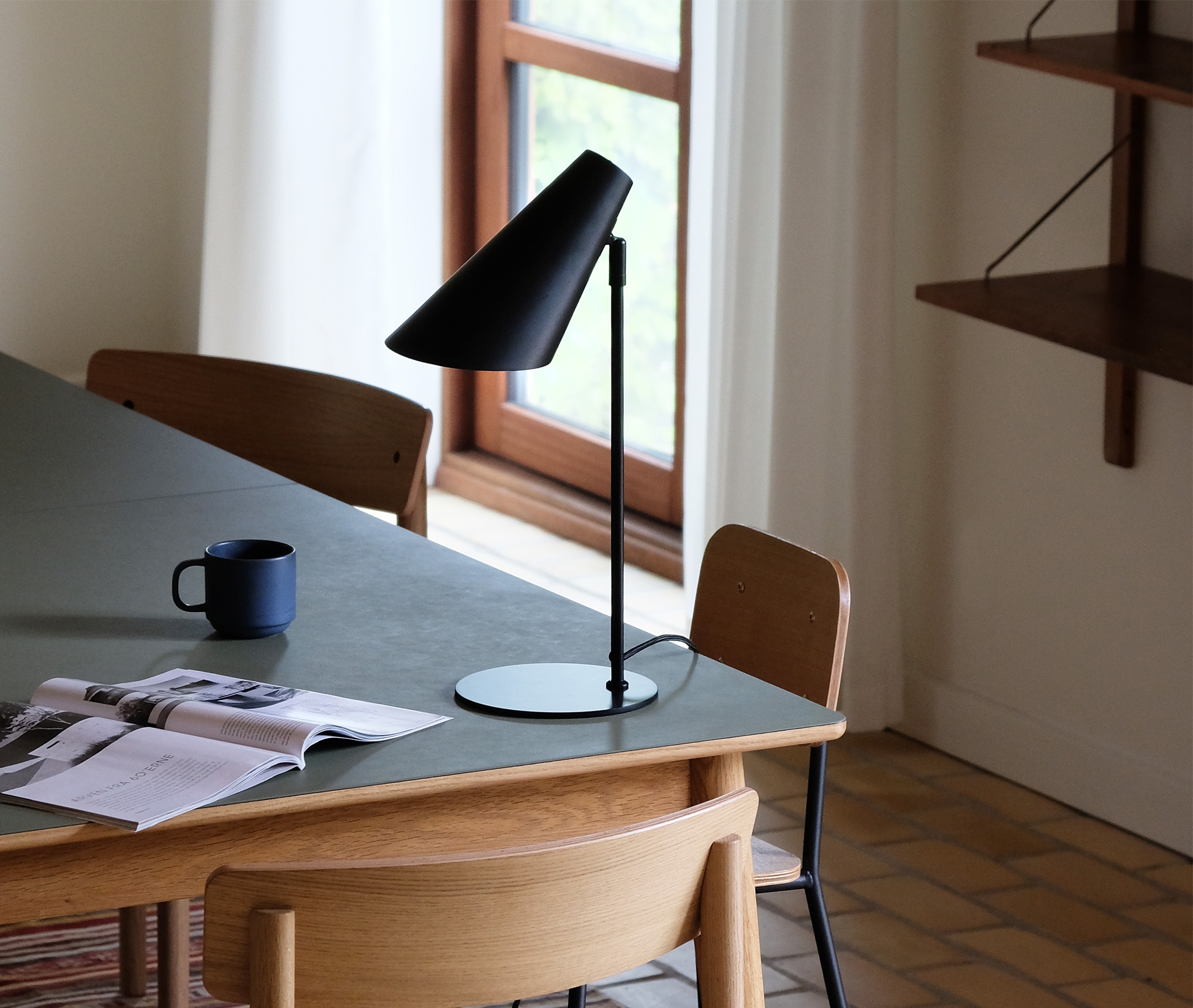 Ljust matrum med trästolar, grönt bord, svart bordslampa, kaffekopp och uppslagen tidning.