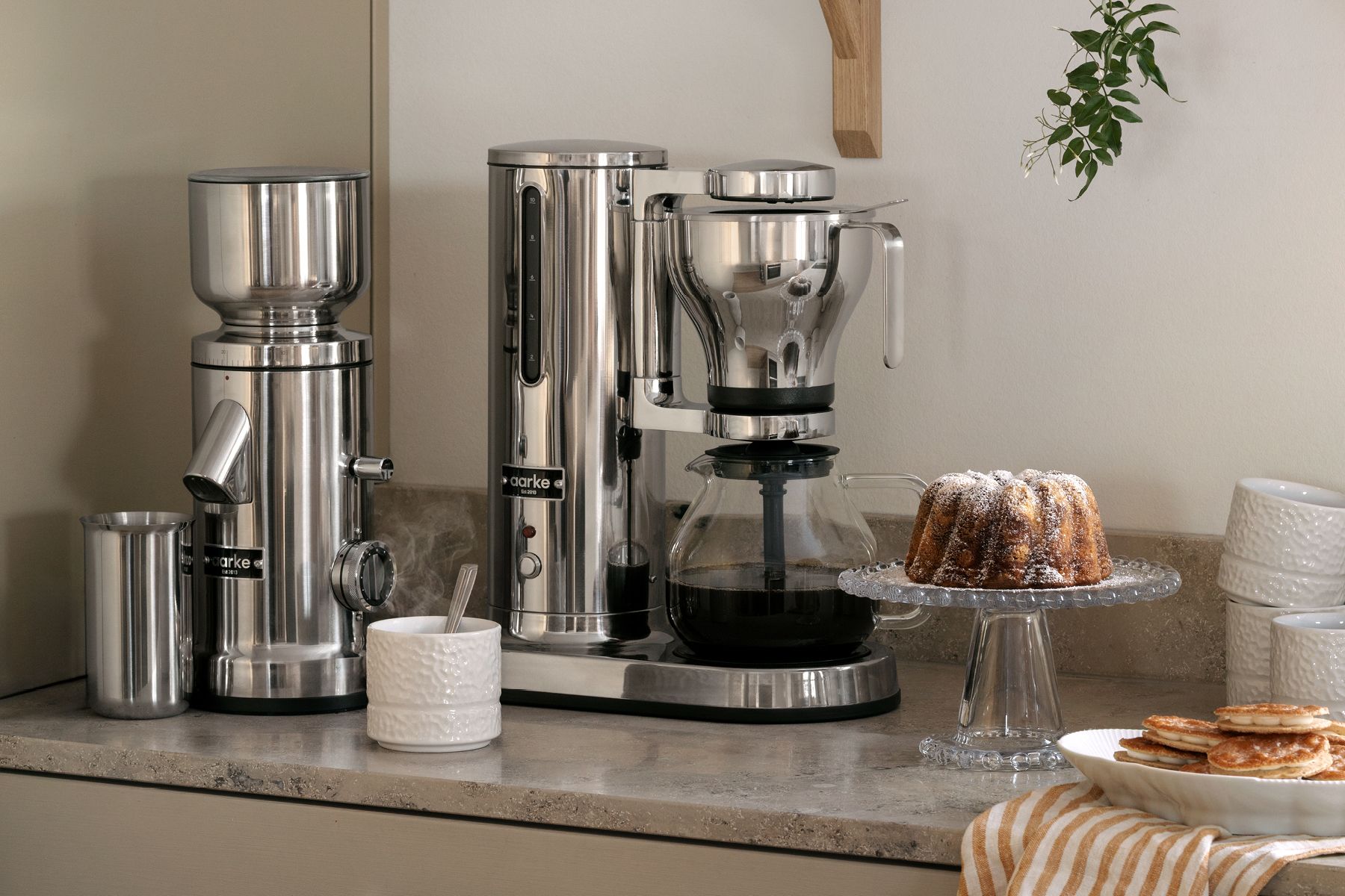 Aarke kaffekvarn och kaffebryggare i rostfritt stål på en köksbänk med en bundtkaka och pannkakor.
