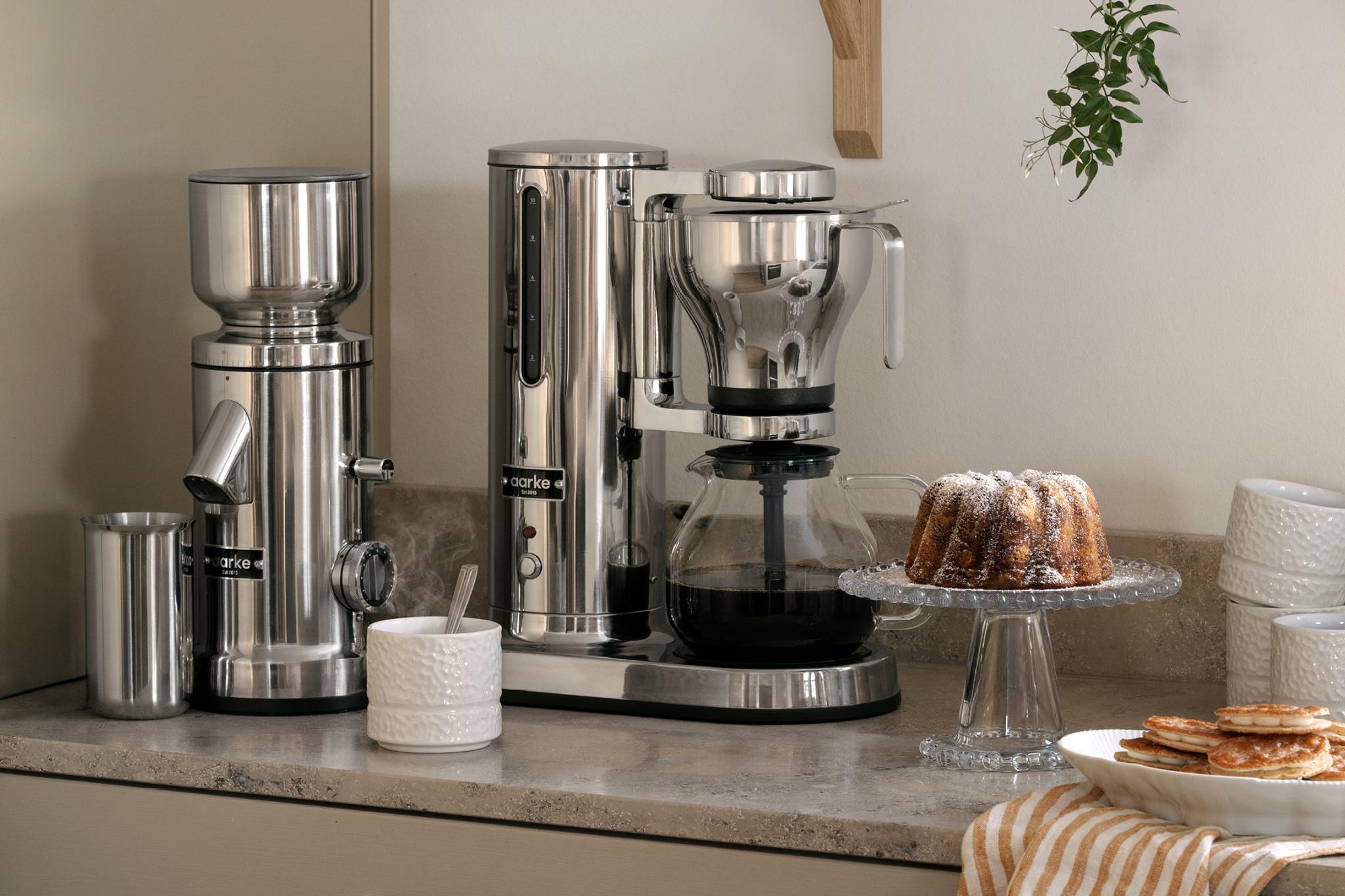 Aarke kaffetrakter, kvern og kaffekaraffel ved siden av en bundtkake på en kjøkkenbenk.