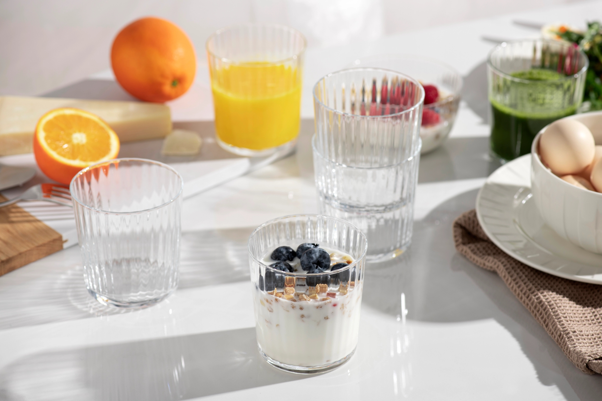 Ljust frukostbord med yoghurt och blåbär, apelsinjuice, grön juice, ägg och färska apelsiner.