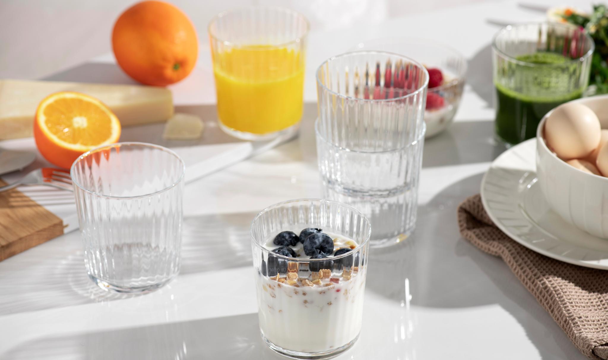 Ljust frukostbord med yoghurt och blåbär, apelsinjuice, grön juice, ägg och färska apelsiner.