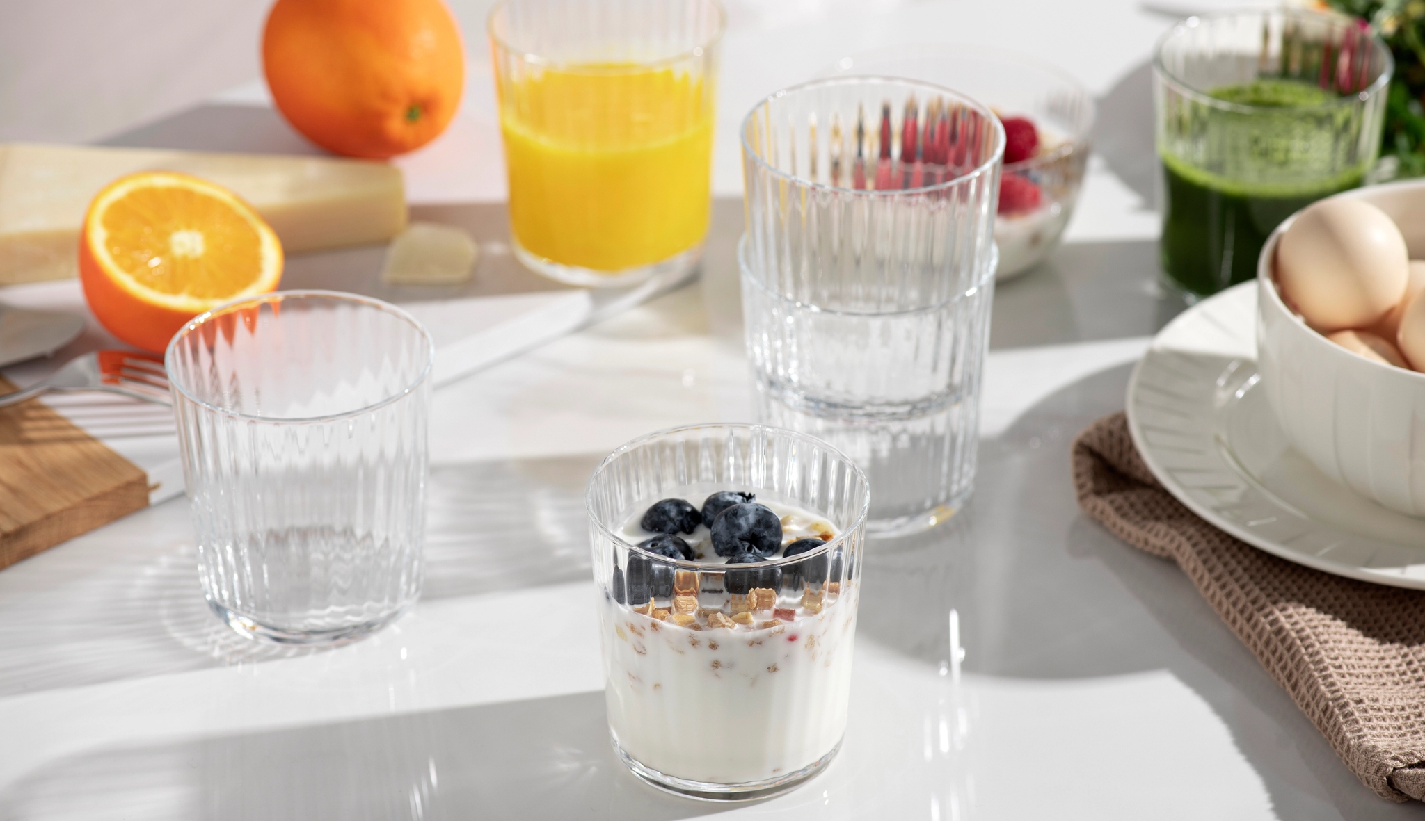 Ljust frukostbord med yoghurt och blåbär, apelsinjuice, grön juice, ägg och färska apelsiner.