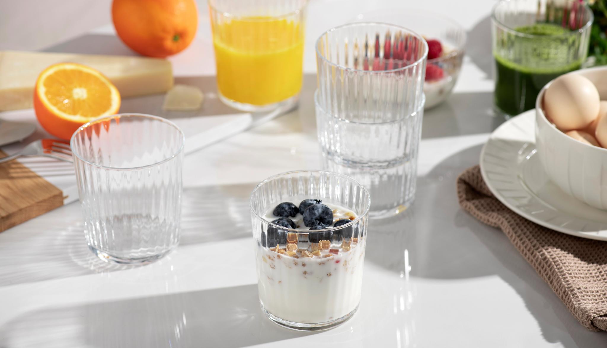 Ljust frukostbord med yoghurt och blåbär, apelsinjuice, grön juice, ägg och färska apelsiner.