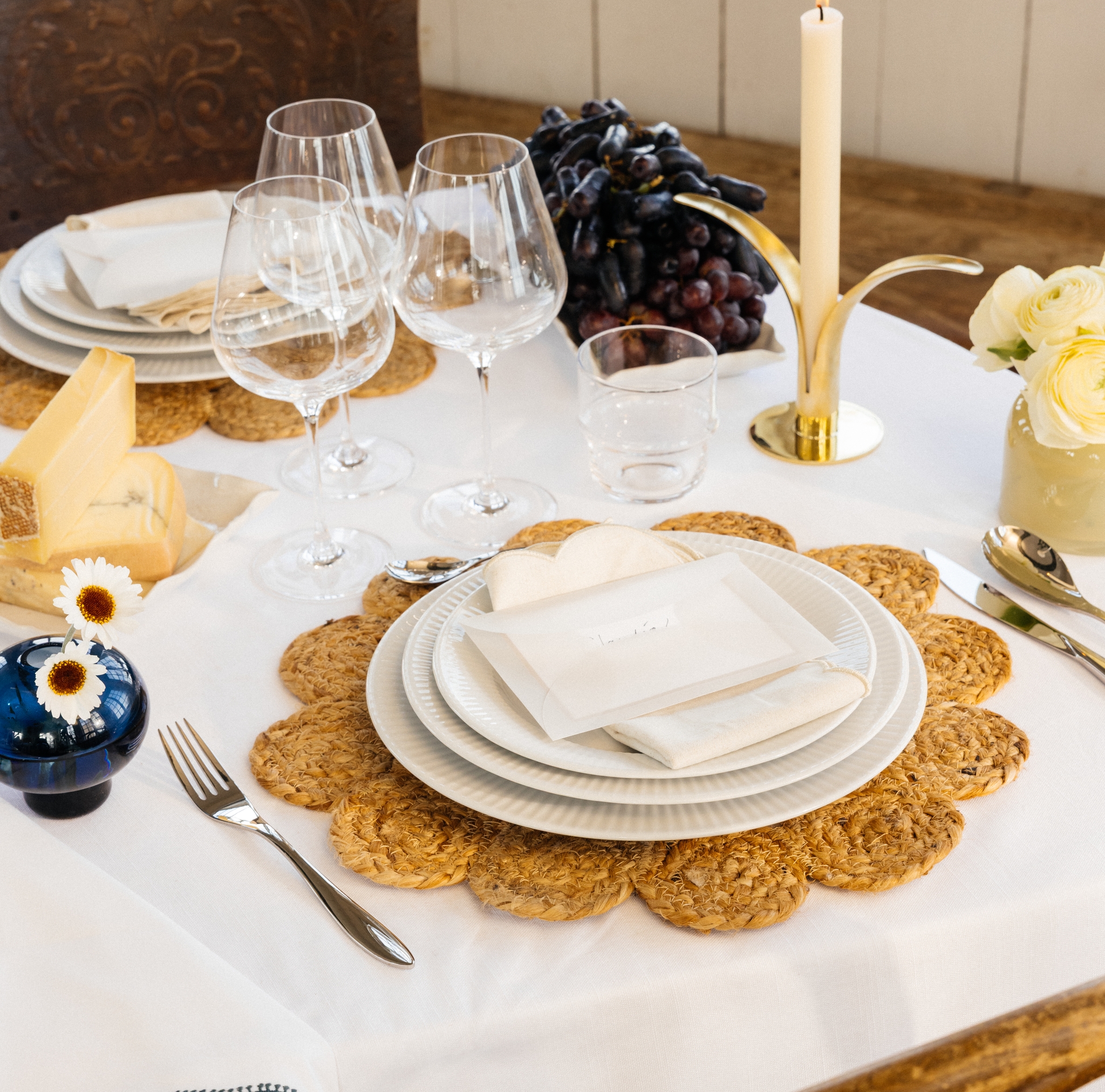 Juhlallinen kattaus, jossa valkoinen pöytäliina, pöytämatot, valkoinen posliini ja keltaiset ja siniset kukkamaljakot