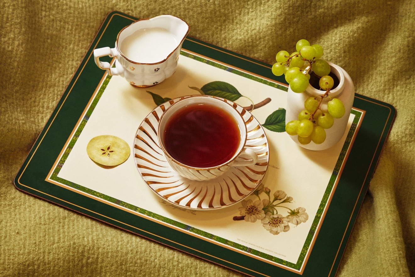 En kopp te, en mjölkkanna, gröna vindruvor och en äppelskiva på en botanisk bordstablett, placerade på en grön filt.