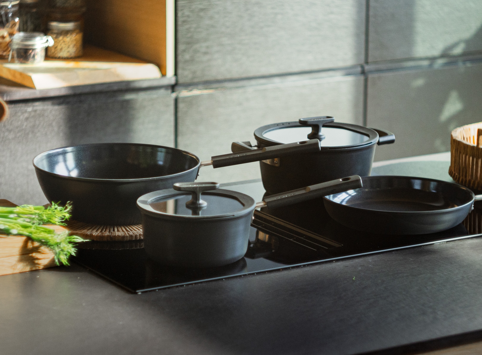 Svarta köksredskap på en modern induktionshäll, med färska grönsaker och burkar i bakgrunden.