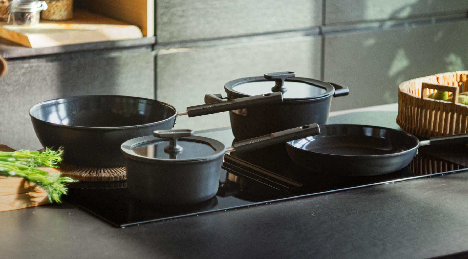 Svarta köksredskap på en modern induktionshäll, med färska grönsaker och burkar i bakgrunden.